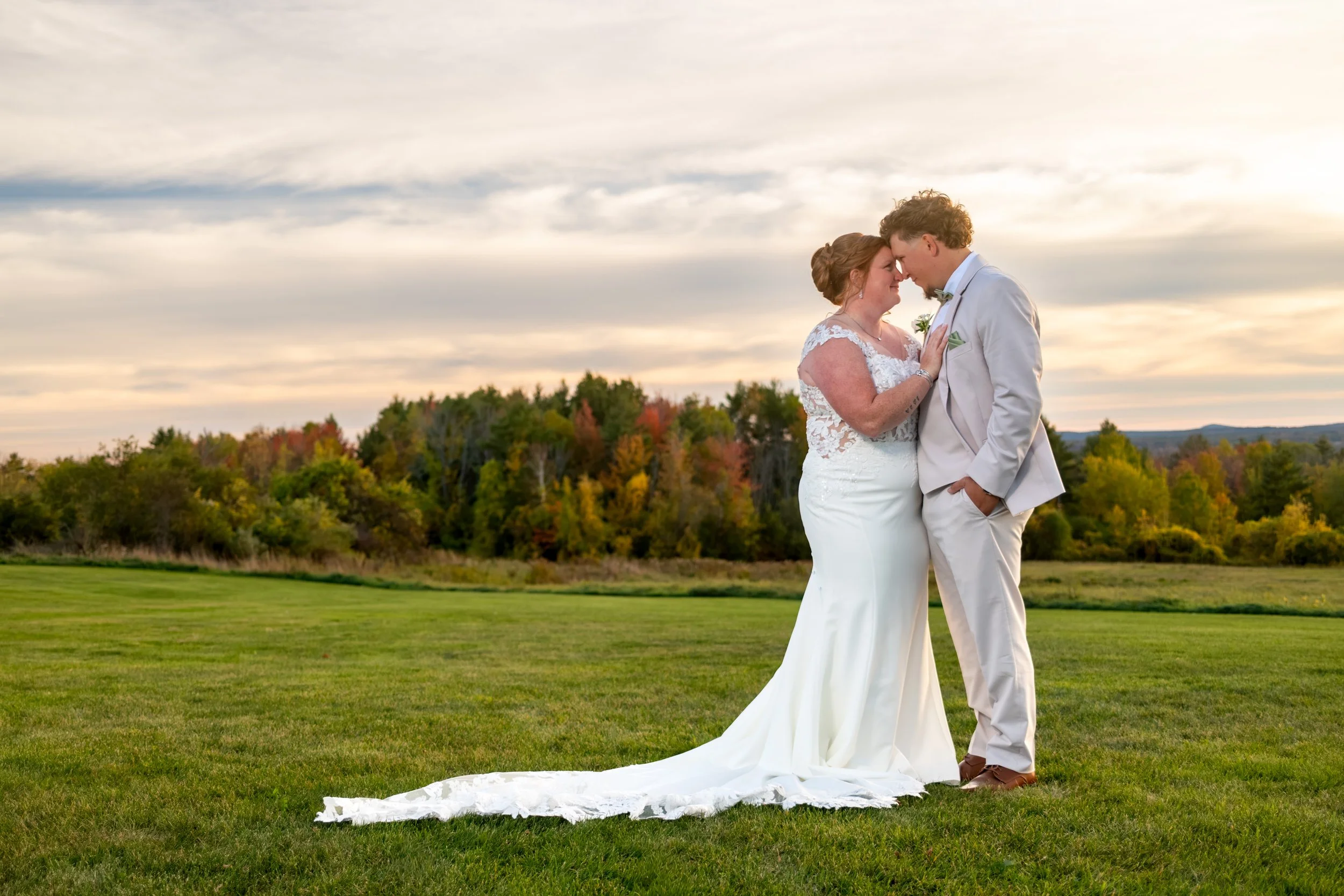 Erika and Andrew kissing in a green field at golden hour with her long train across the grass and fall foliage behind them at the Maine Wedding Barn and Event Center in Minot Maine fall wedding photography VMG Productions