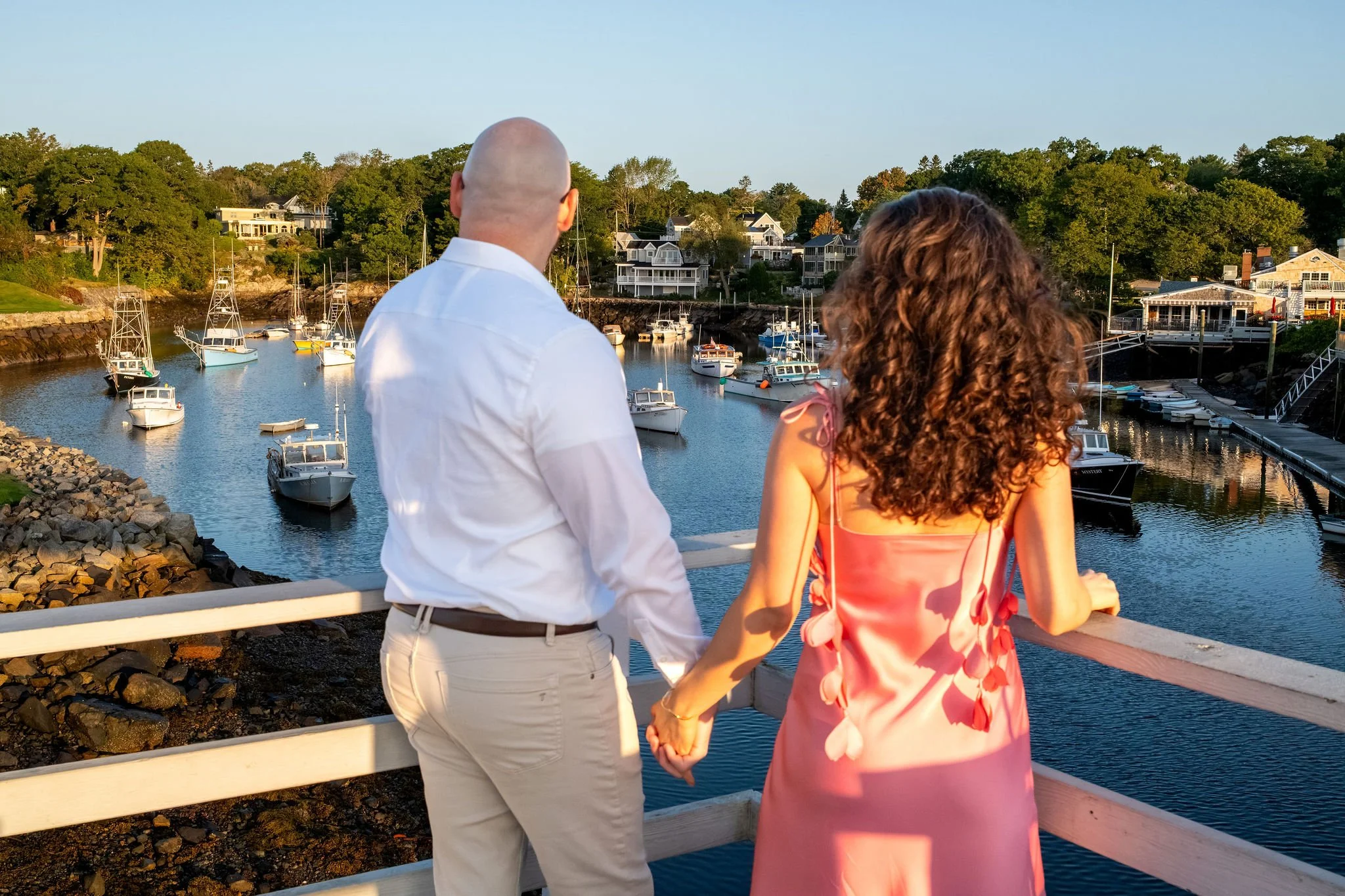 Zack and Helena holding hands at the railing and looking out at the boats in Perkins Cove on Marginal Way in Ogunquit Maine sunrise proposal and engagement photography VMG Productions