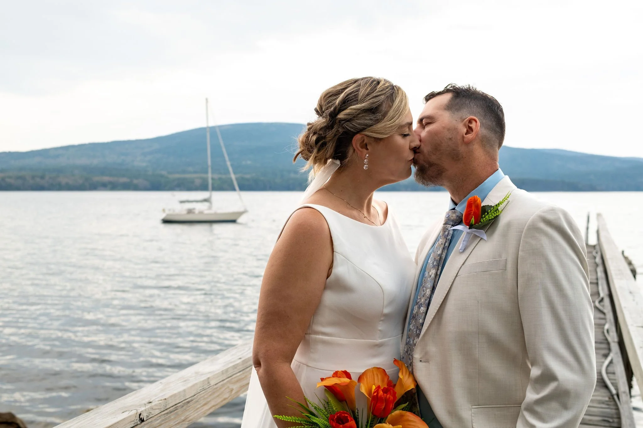 Alli and Adam Baudoin kissing on the wooden dock with a sailboat on Somes Sound behind them at their private residence spring wedding in Mount Desert Maine wedding photography VMG Productions