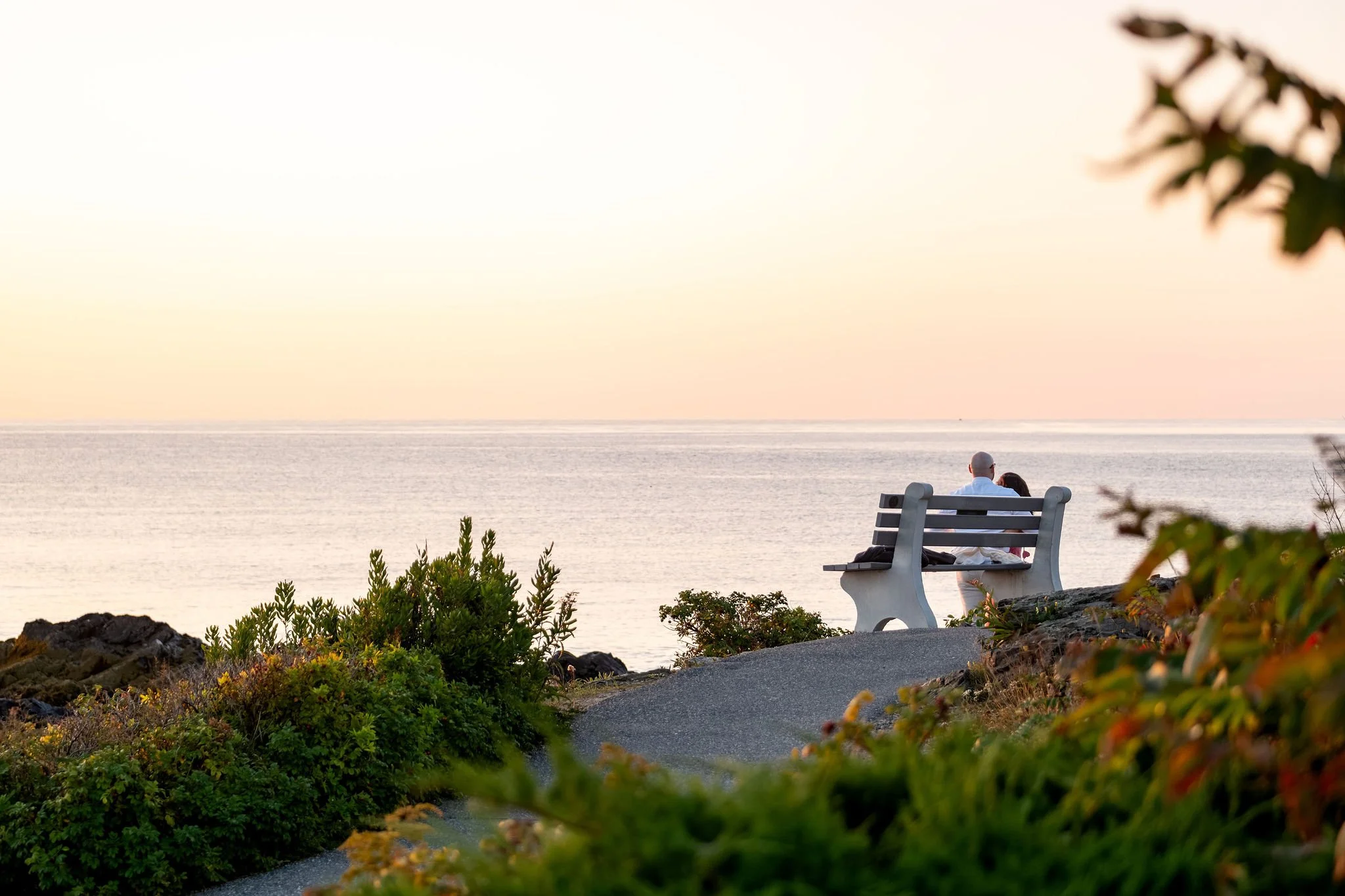 Zack and Helena sitting on a bench at the edge of the path looking out at the sunrise over the ocean on Marginal Way in Ogunquit Maine sunrise proposal and engagement photography VMG Productions