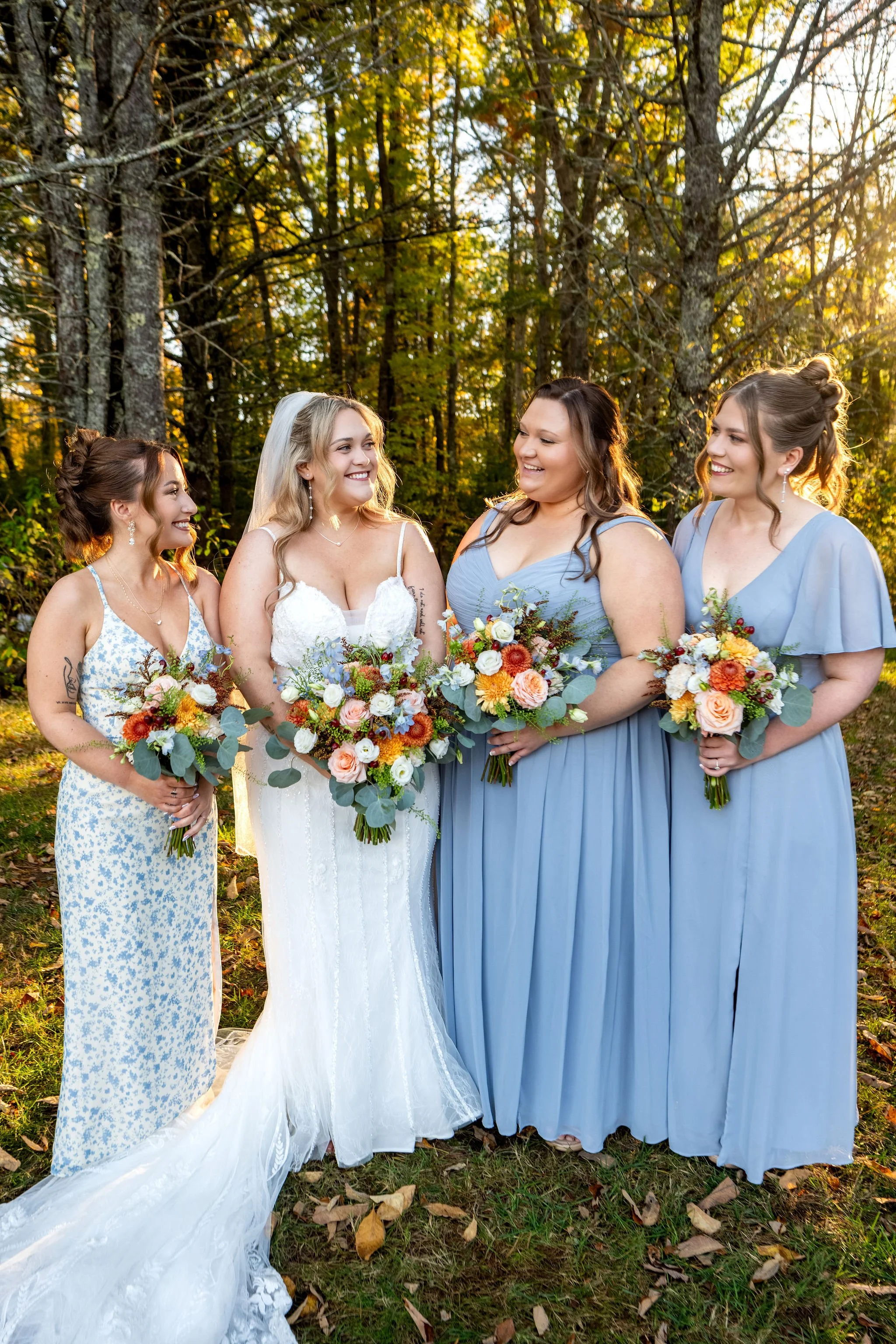 Ellie and her bridesmaids laughing together holding their bouquets in the forest light at Boothbay Playhouse in Boothbay Maine fall wedding photography VMG Productions
