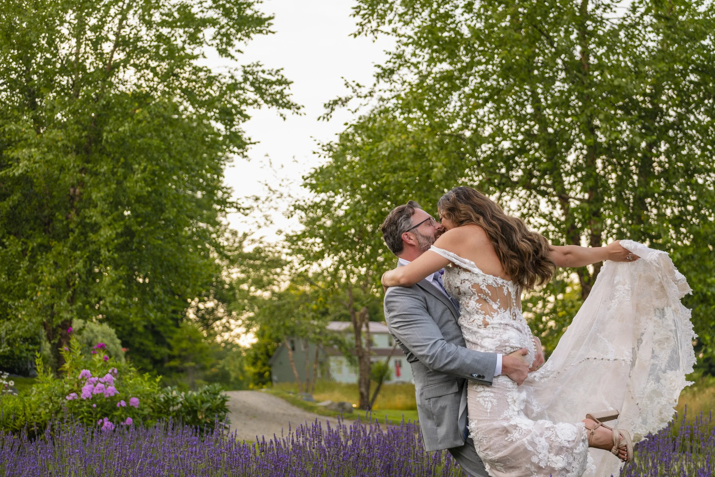 Groom lifting bride in off-shoulder lace wedding gown for a kiss in lavender field at golden hour with trees and warm summer light in background at Marianmade Farm Wiscasset Maine summer wedding 2024
