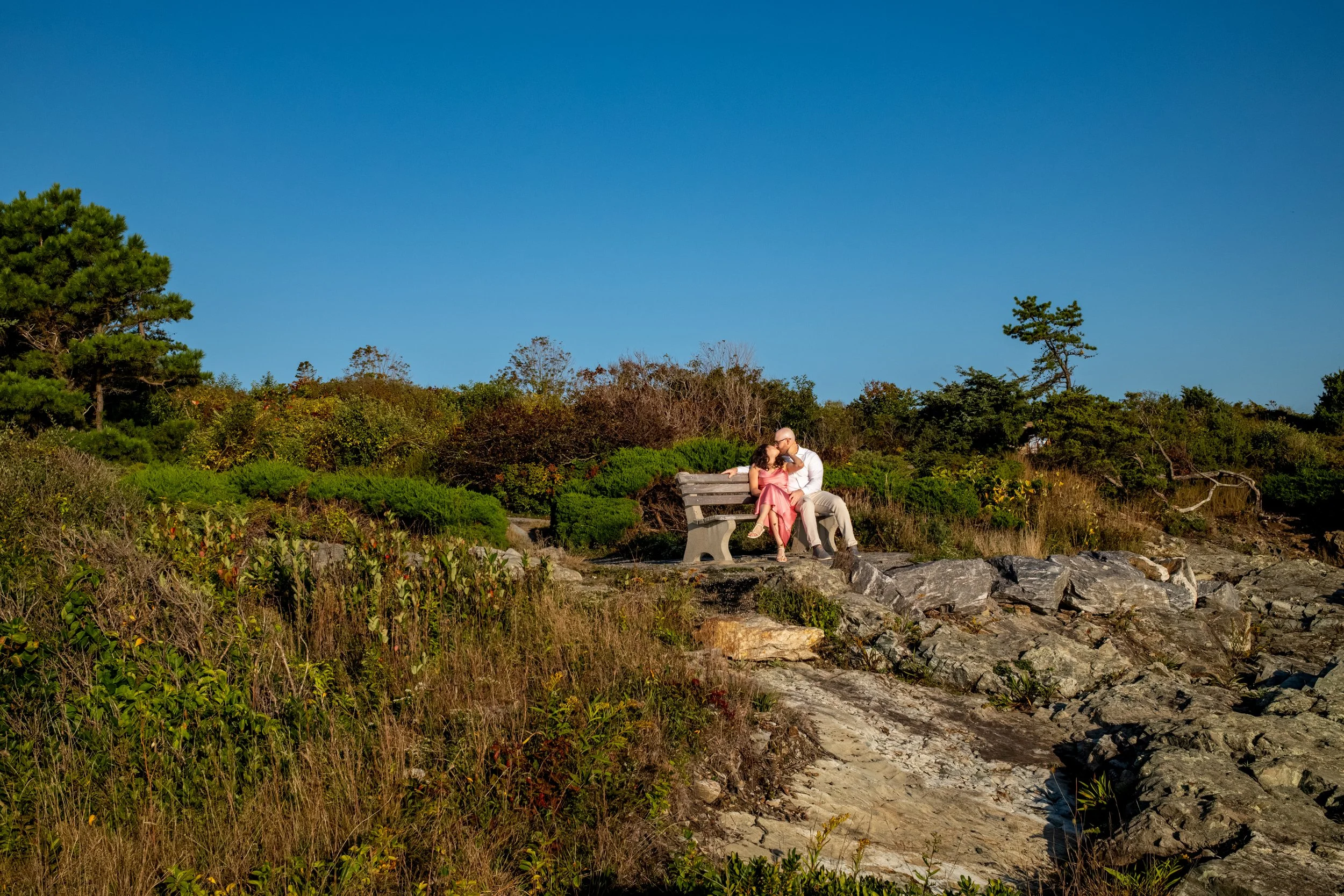 Zack and Helena sitting on a bench in the distance along the rocky Marginal Way path under a deep blue sky on Marginal Way in Ogunquit Maine sunrise proposal and engagement photography VMG Productions