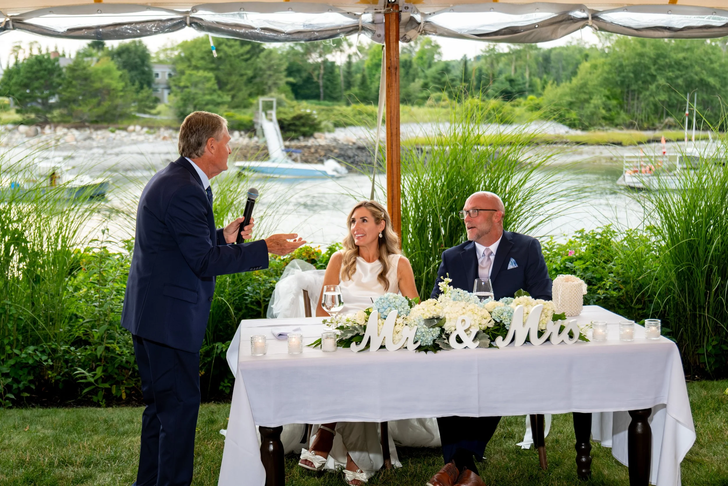 Ashley and Marc Draper at the Mr and Mrs sweetheart table dressed in hydrangeas during a toast at The Breakwater Inn in Kennebunkport Maine summer wedding photography VMG Productions