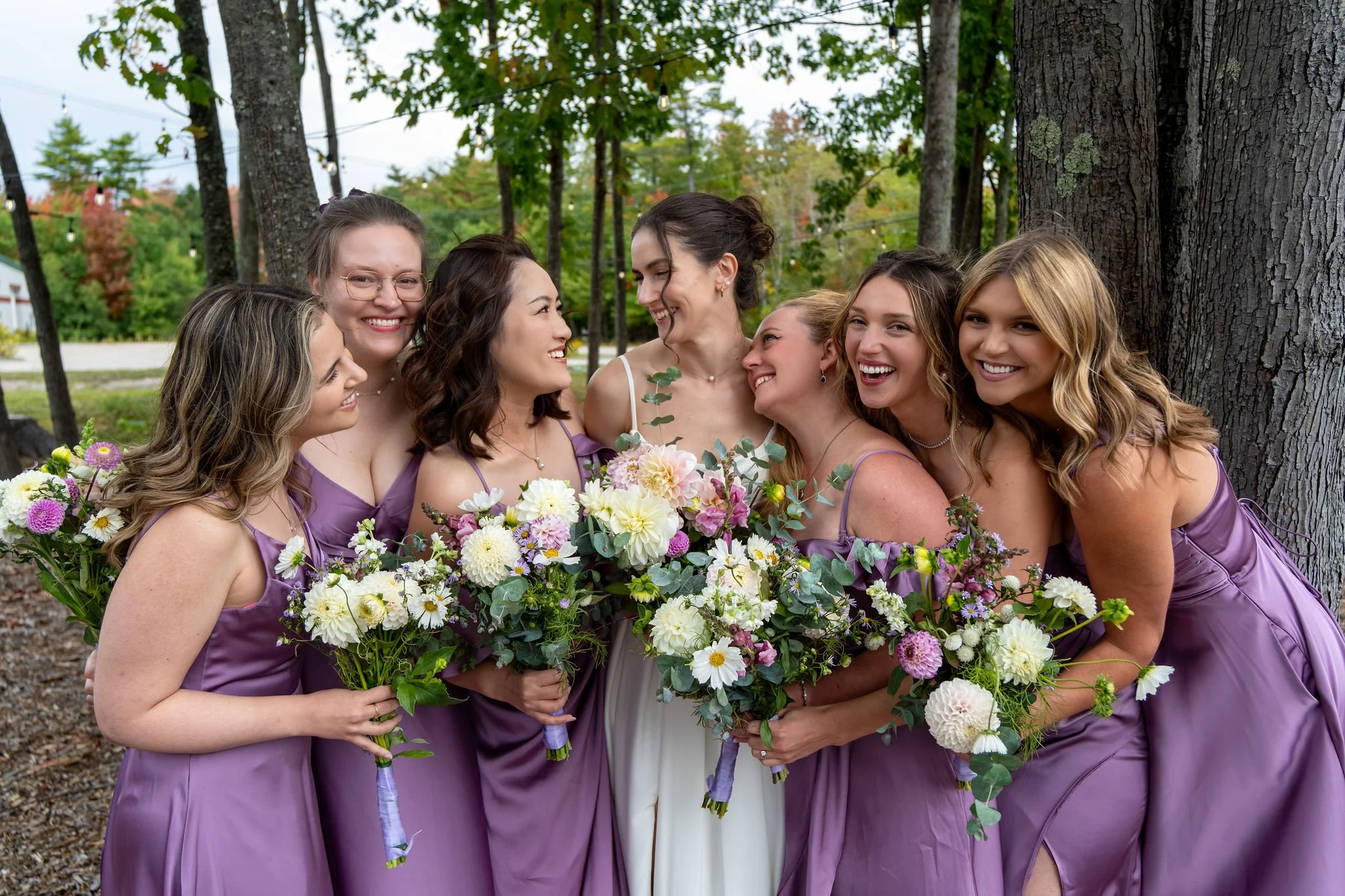 Victoria laughing with her bridesmaids in lavender satin dresses holding wildflower bouquets at The Cellars at Allagash Brewing Company in Portland Maine fall wedding photography VMG Productions