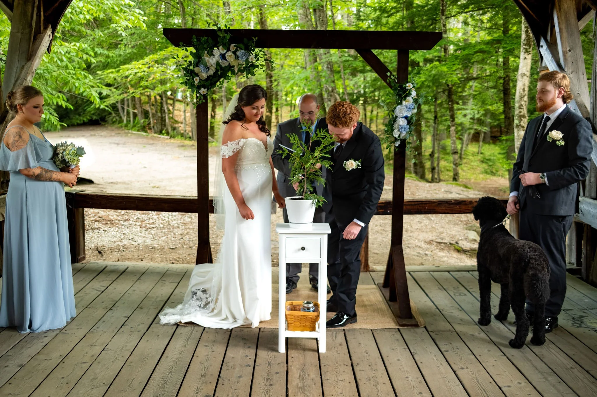 Alyssa and Colby planting a tree together during their garden unity ceremony beneath the floral arbor with Ryder watching at Sunday River Covered Bridge Newry Maine summer wedding photography VMG Productions