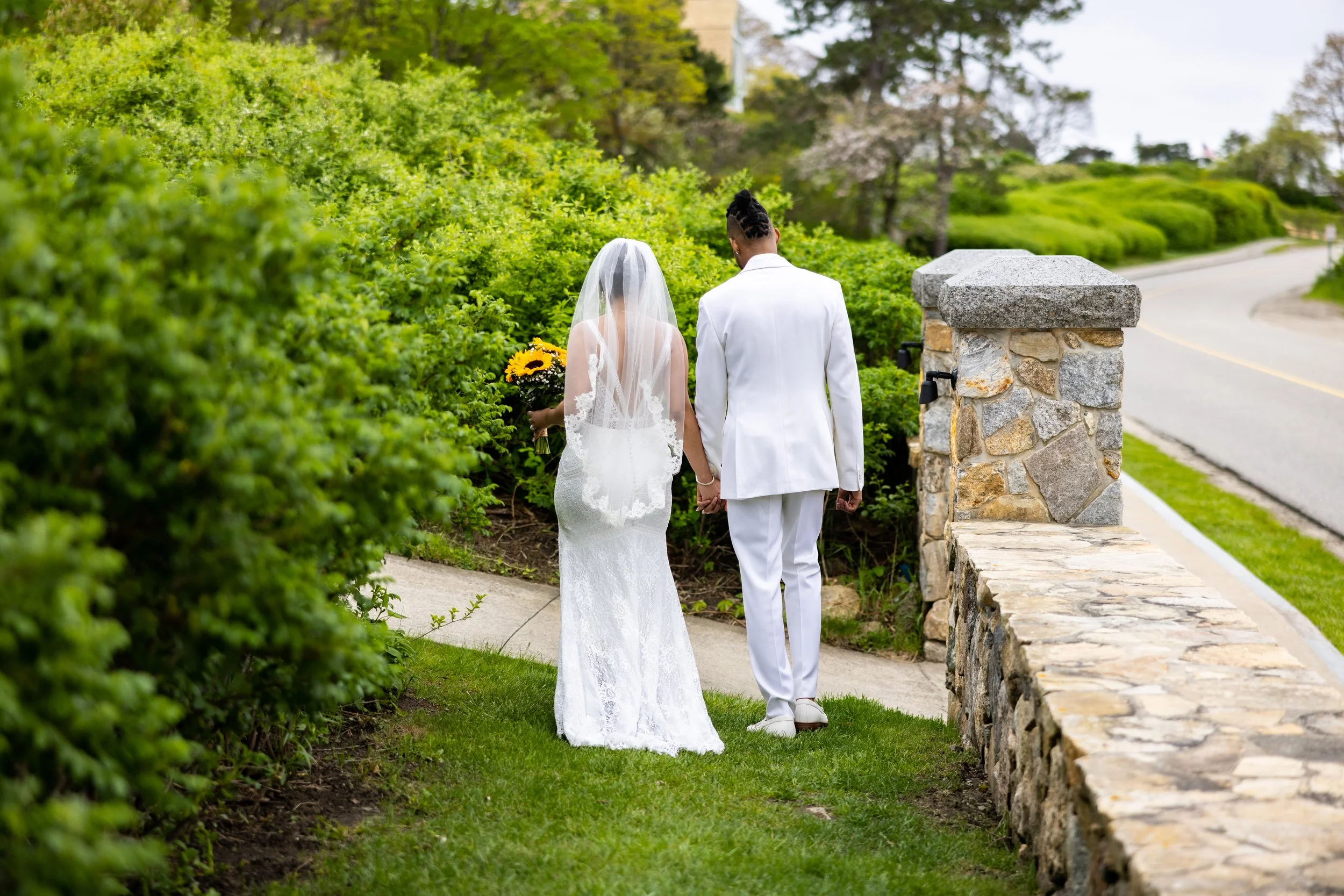 Bride in lace wedding gown with long cathedral veil holding sunflower bouquet walking hand in hand with groom in white suit on stone pathway surrounded by lush green hedges at The Colony Hotel Kennebunkport Maine May 2022
