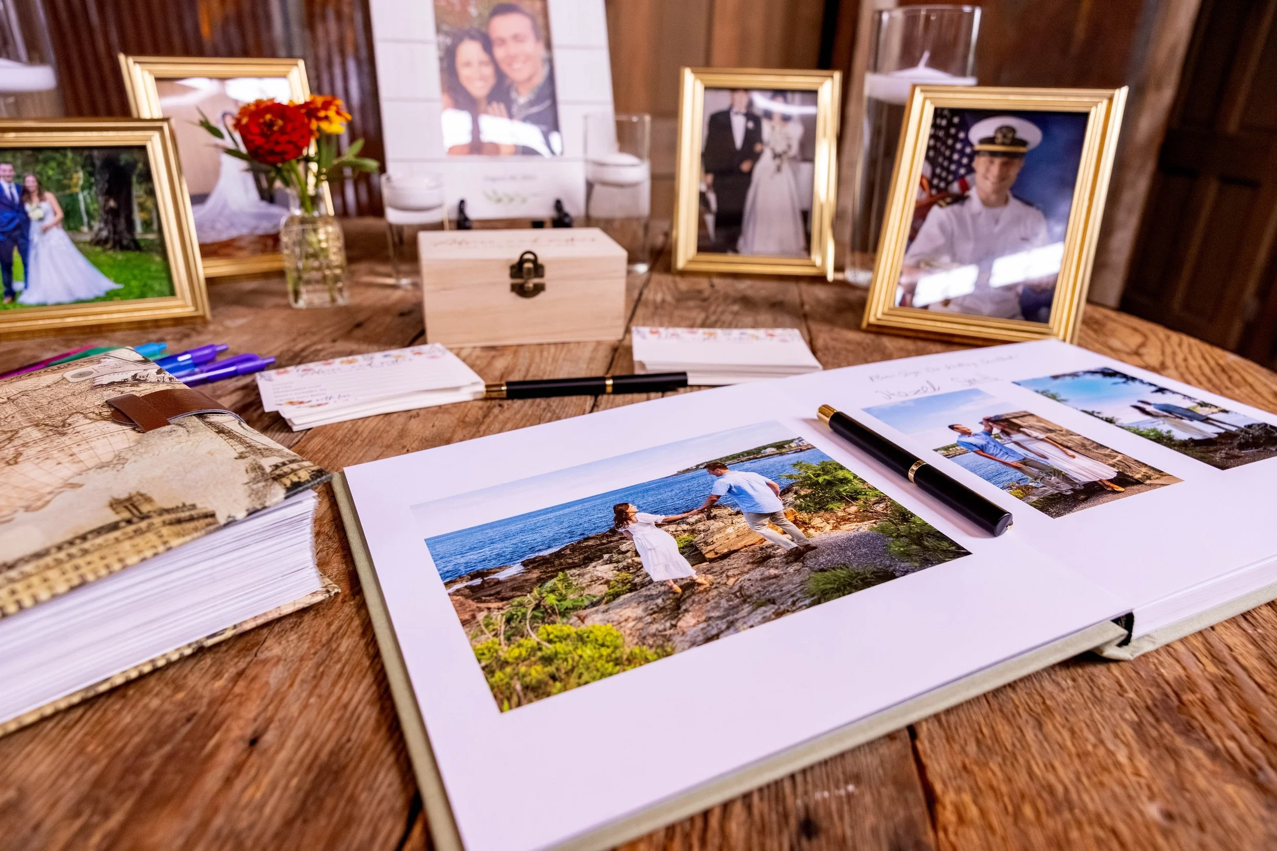 Wedding guest book table at Mountain Star Estate Rangeley Maine barn wedding 2025 with printed engagement photo album open showing coastal photos framed family photographs gold frames wildflower bouquet wooden keepsake chest and advice cards with pens