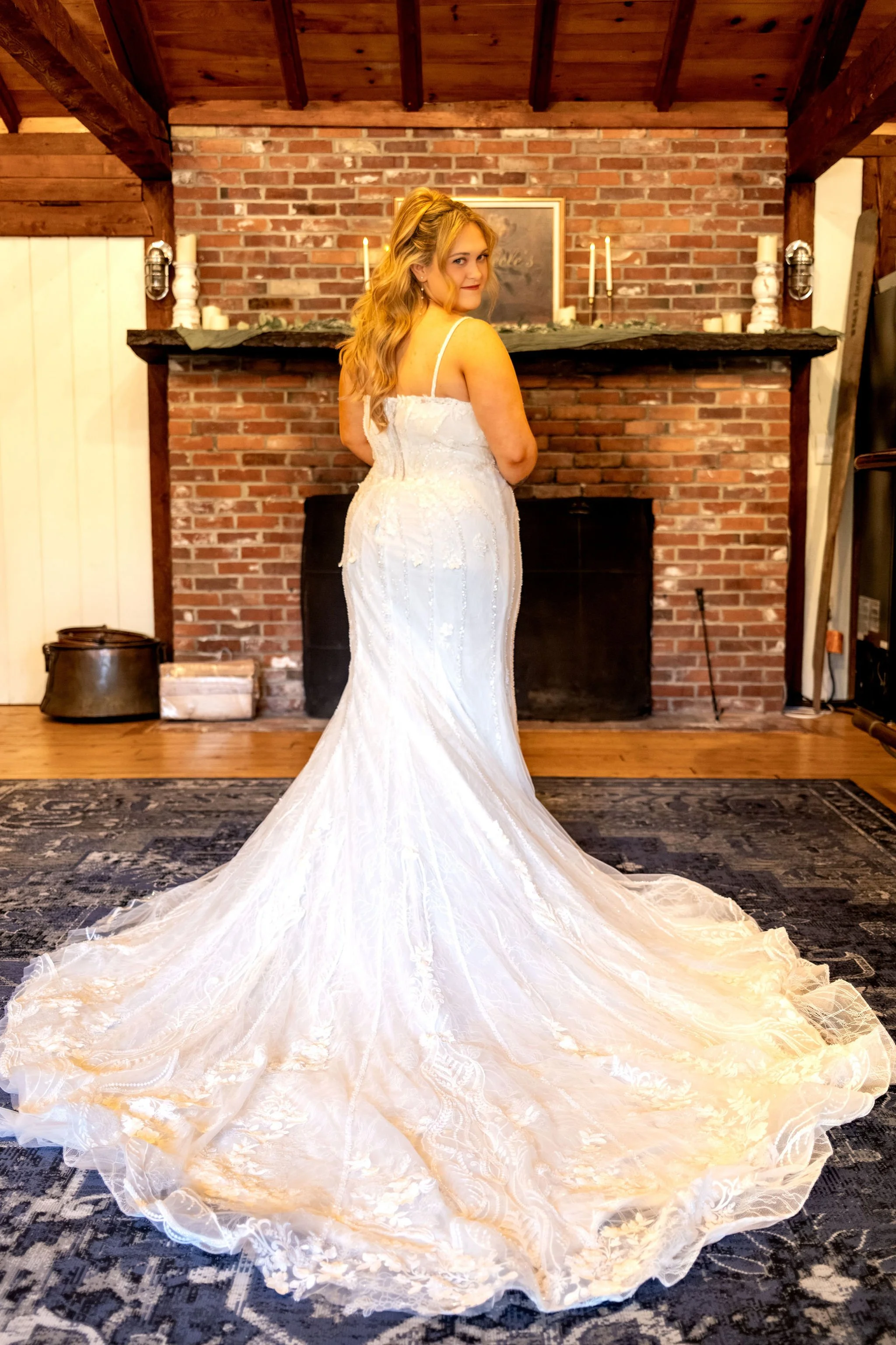 Ellie looking back over her shoulder in her gown with the long train spread across the rug in front of the brick fireplace at Boothbay Playhouse in Boothbay Maine fall wedding photography VMG Productions