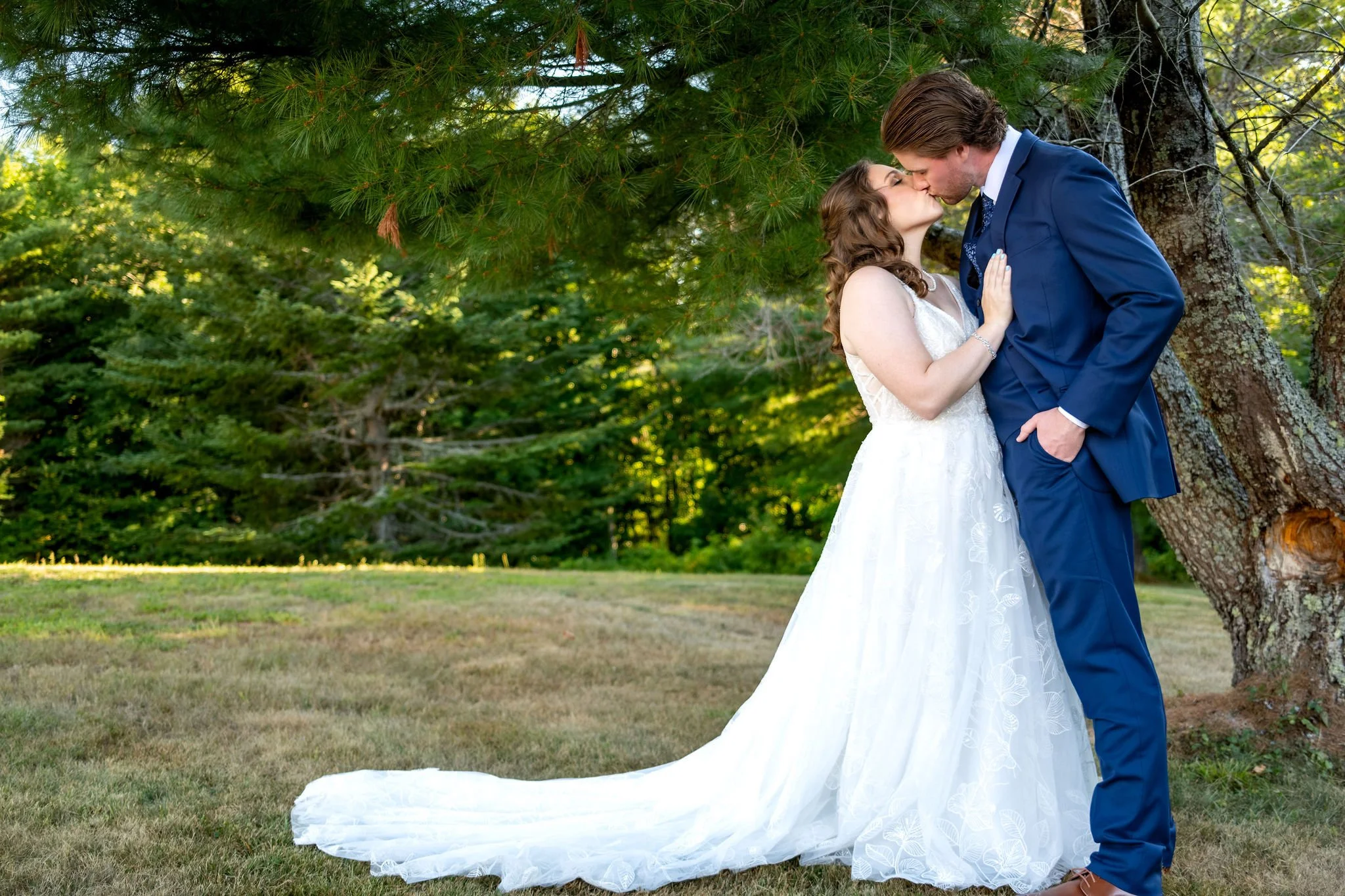 Zachary and Hollie kissing beneath the pine boughs at the edge of the woods at The Lucerne Inn in Dedham Maine summer wedding photography VMG Productions