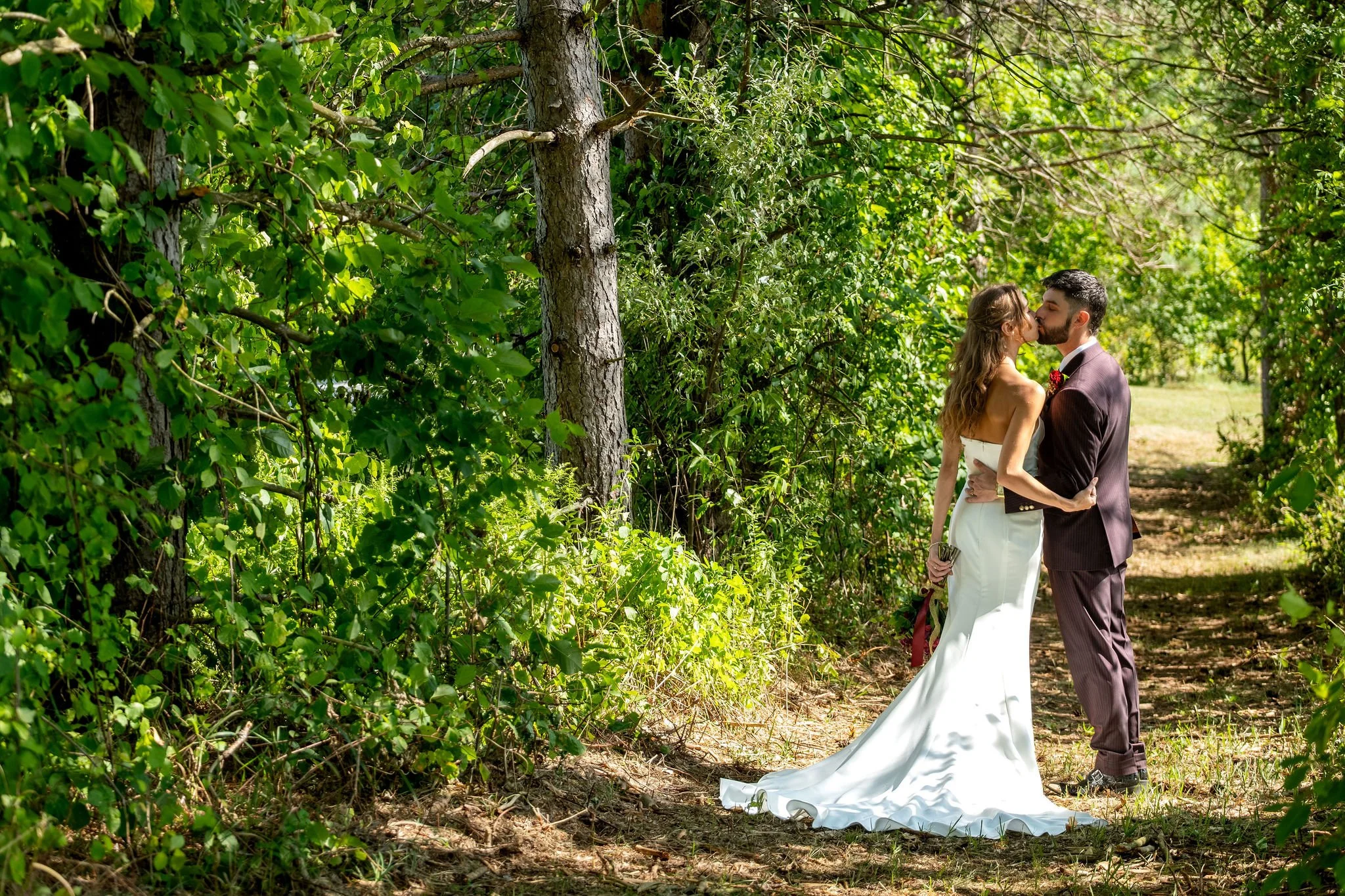 Cassidy and Chase Begin kissing at the edge of a wooded trail with her train on the pine needles at Maple Hill Farm Inn and Conference Center in Hallowell Maine summer wedding photography VMG Productions