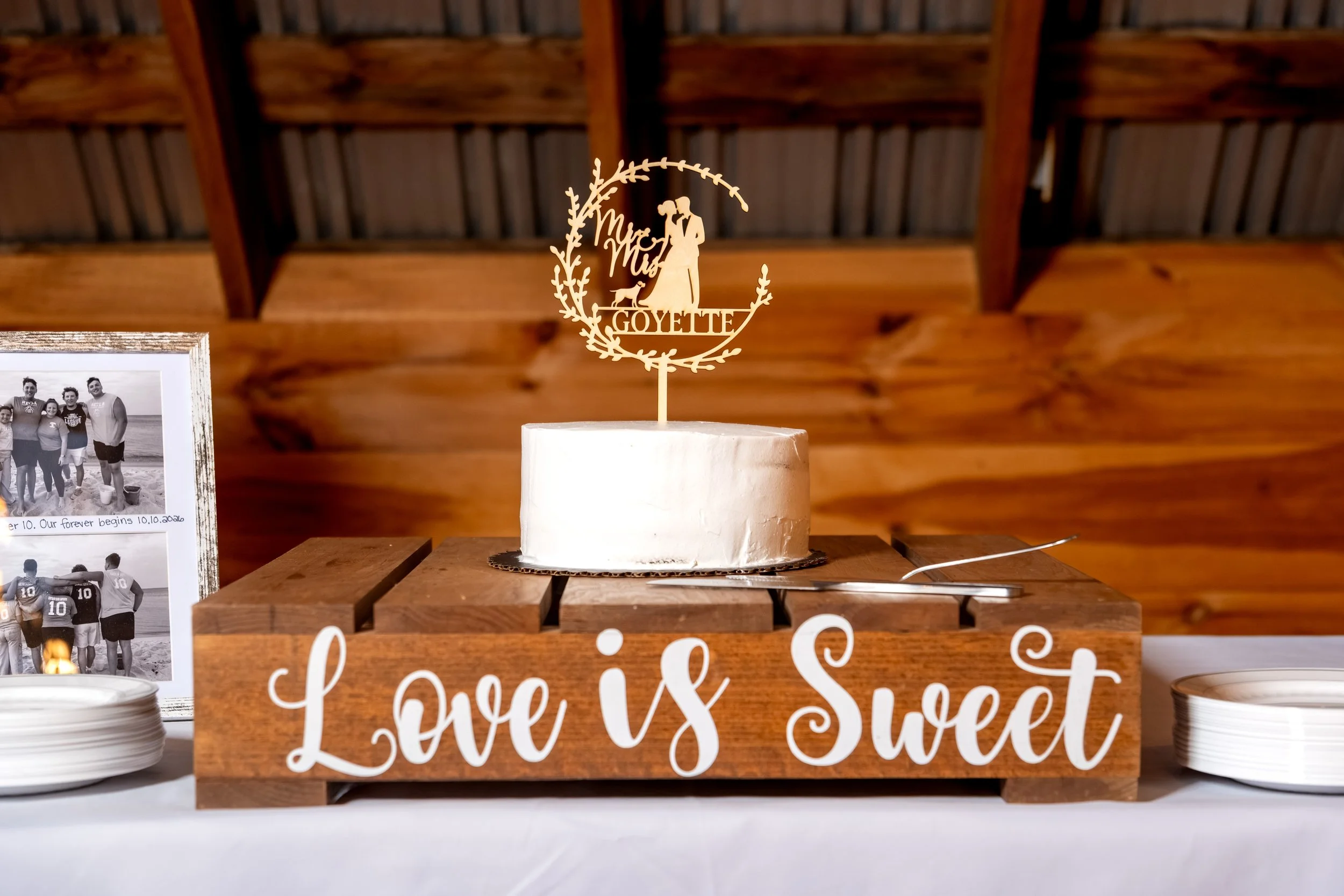 The wedding cake on a wooden stand next to a Love is Sweet sign at the Maine Wedding Barn and Event Center in Minot Maine fall wedding photography VMG Productions