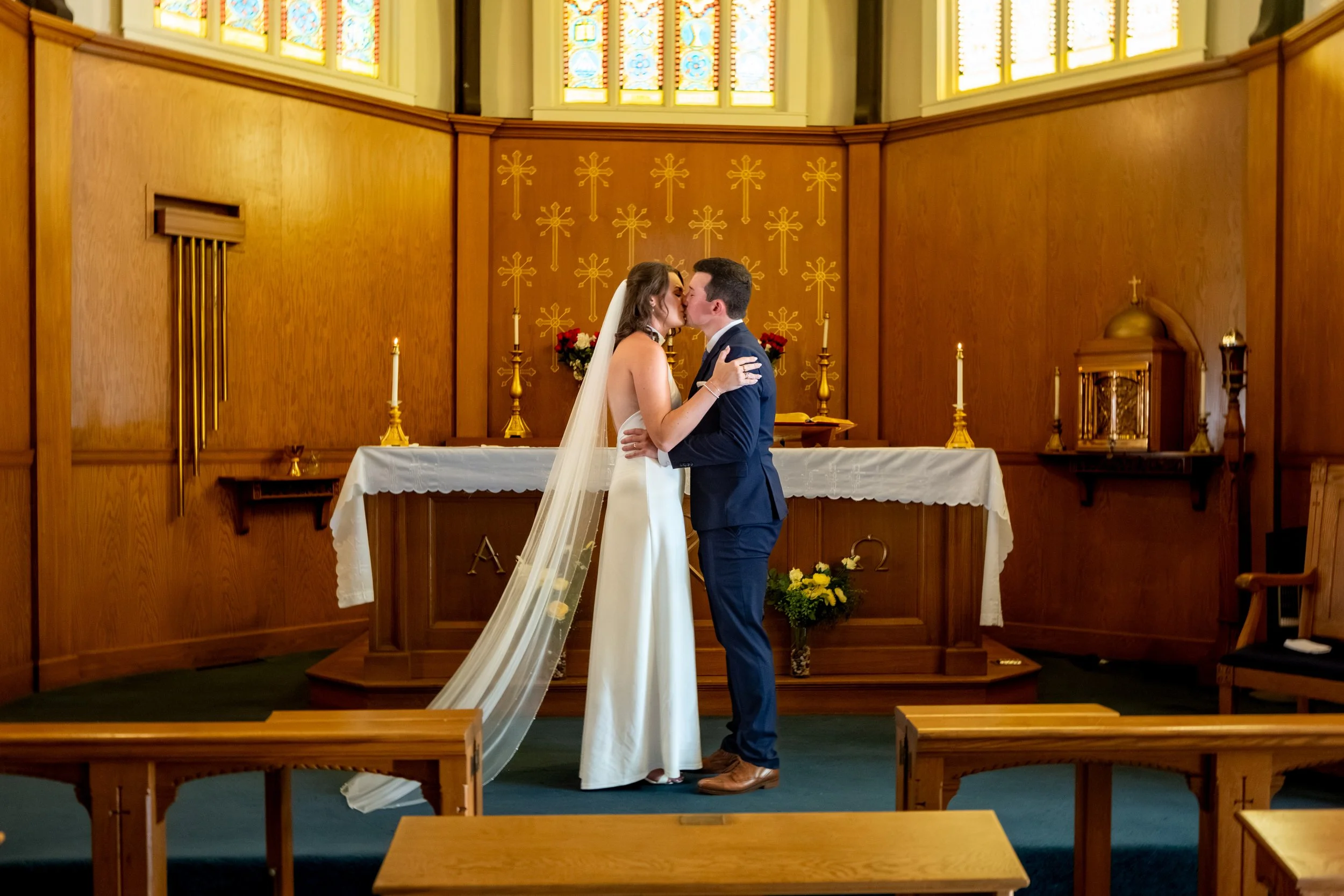 Caroline and Anthony Pirolli sharing their first kiss at the altar beneath the stained glass at Star of the Sea in York Maine wedding photography VMG Productions