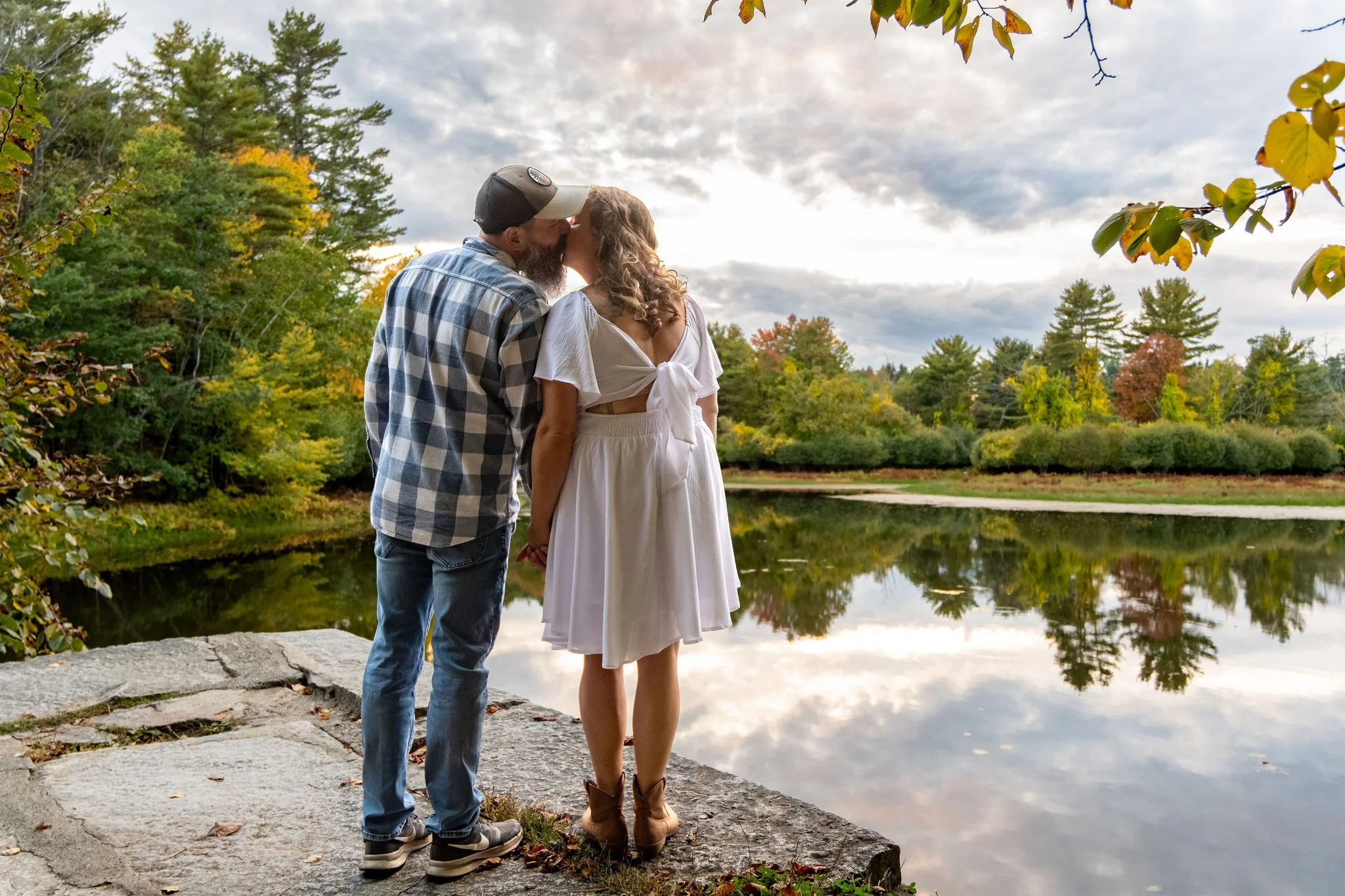Dave kissing Alison on the forehead as they stand at the edge of the pond with the clouds breaking overhead at Vaughan Woods in Hallowell Maine fall engagement photography VMG Productions