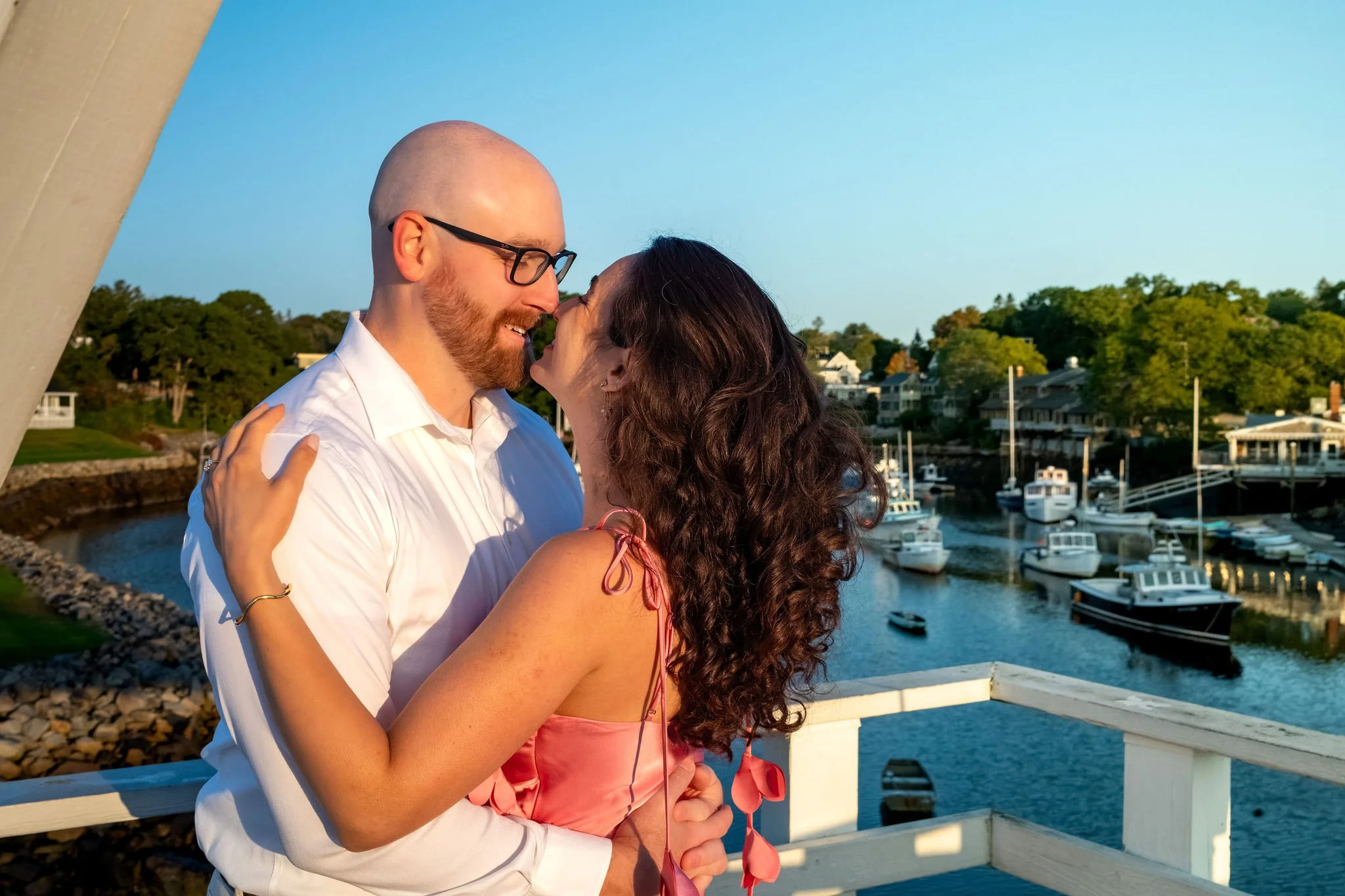Zack and Helena sharing a kiss on the footbridge with moored boats in the harbor behind them on Marginal Way in Ogunquit Maine sunrise proposal and engagement photography VMG Productions