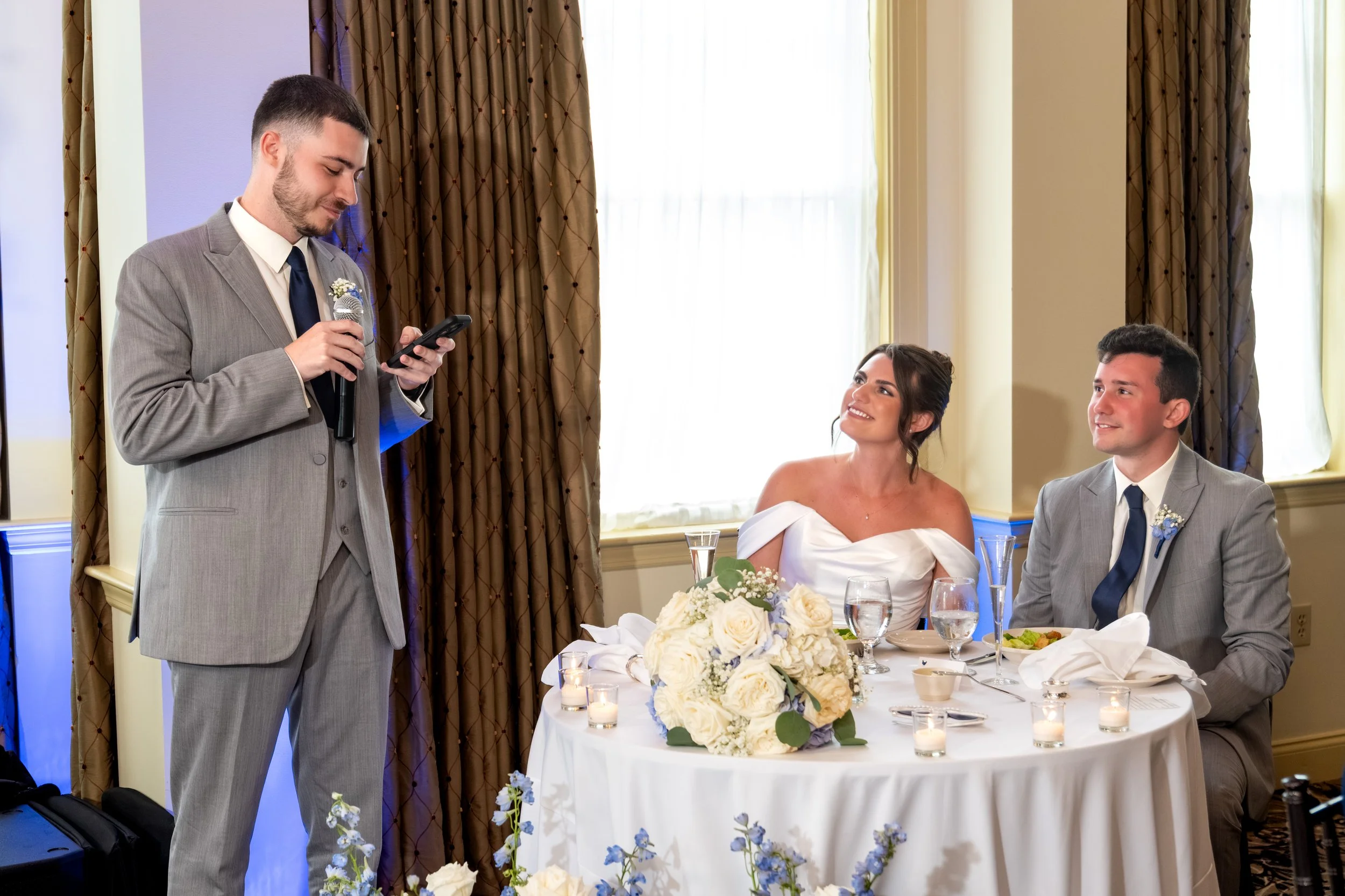 Best man Travis Peabody giving his speech beside the sweetheart table as Caroline and Anthony look up at him at Union Bluff Hotel in York Maine wedding photography VMG Productions