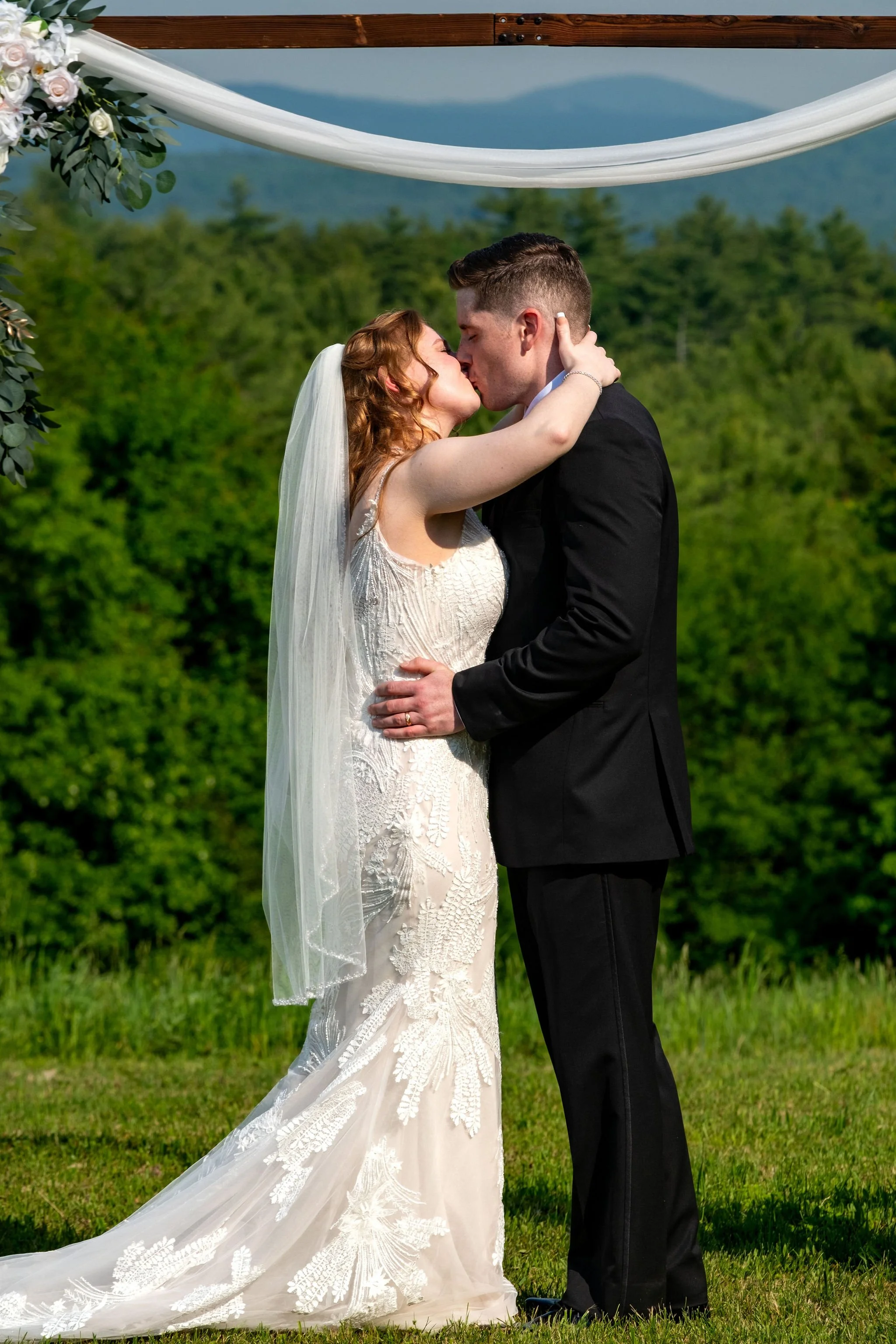 Kayla and Ethan DeAngelis first kiss as a married couple under the arch with the New Hampshire mountains behind them at their Sanbornton backyard wedding photography VMG Productions