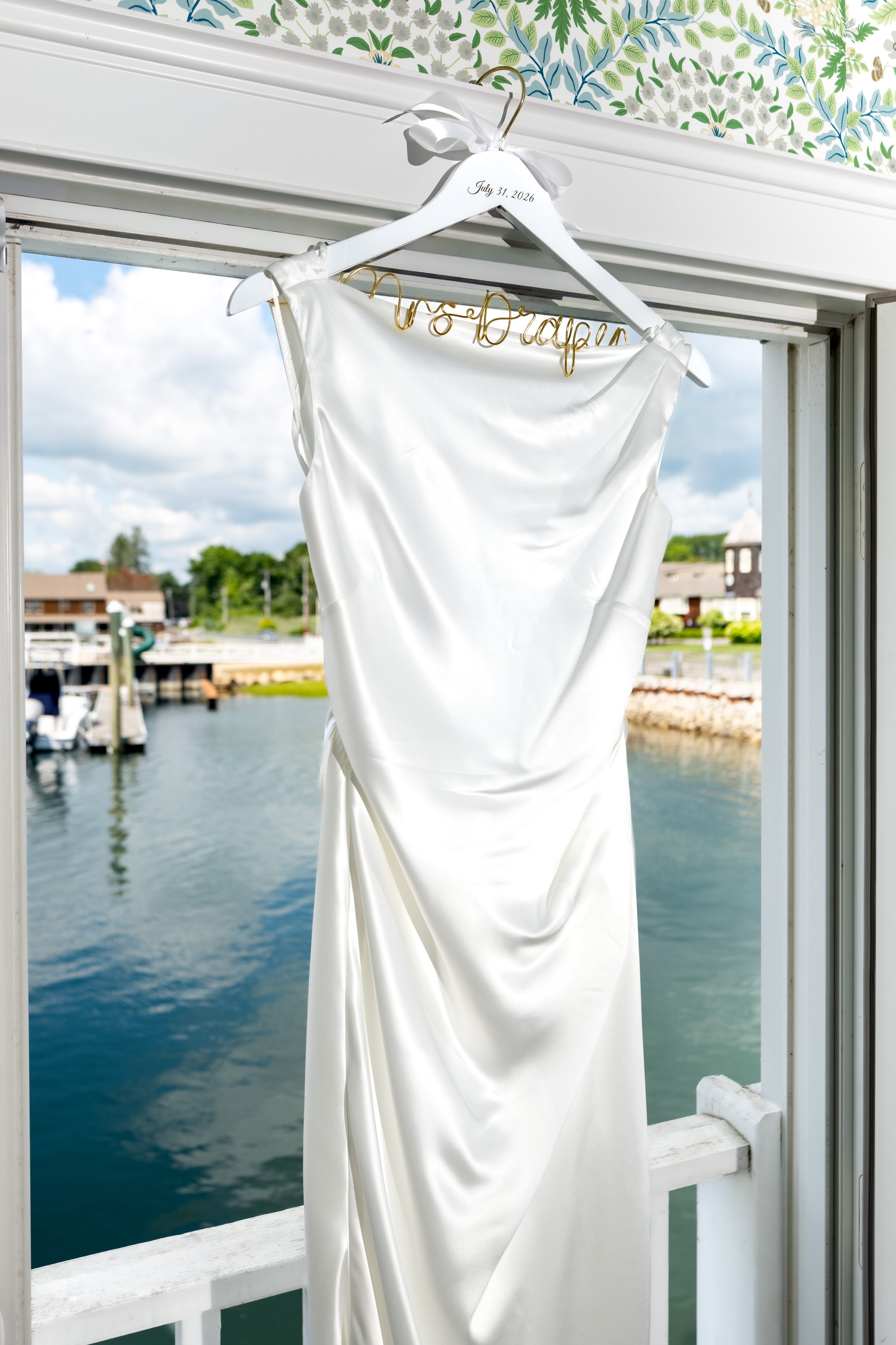 Ashley Draper's wedding gown on a custom Mrs. Draper hanger dated July 31 2026 hanging in the doorway over the harbor at The Breakwater Inn in Kennebunkport Maine summer wedding photography VMG Productions