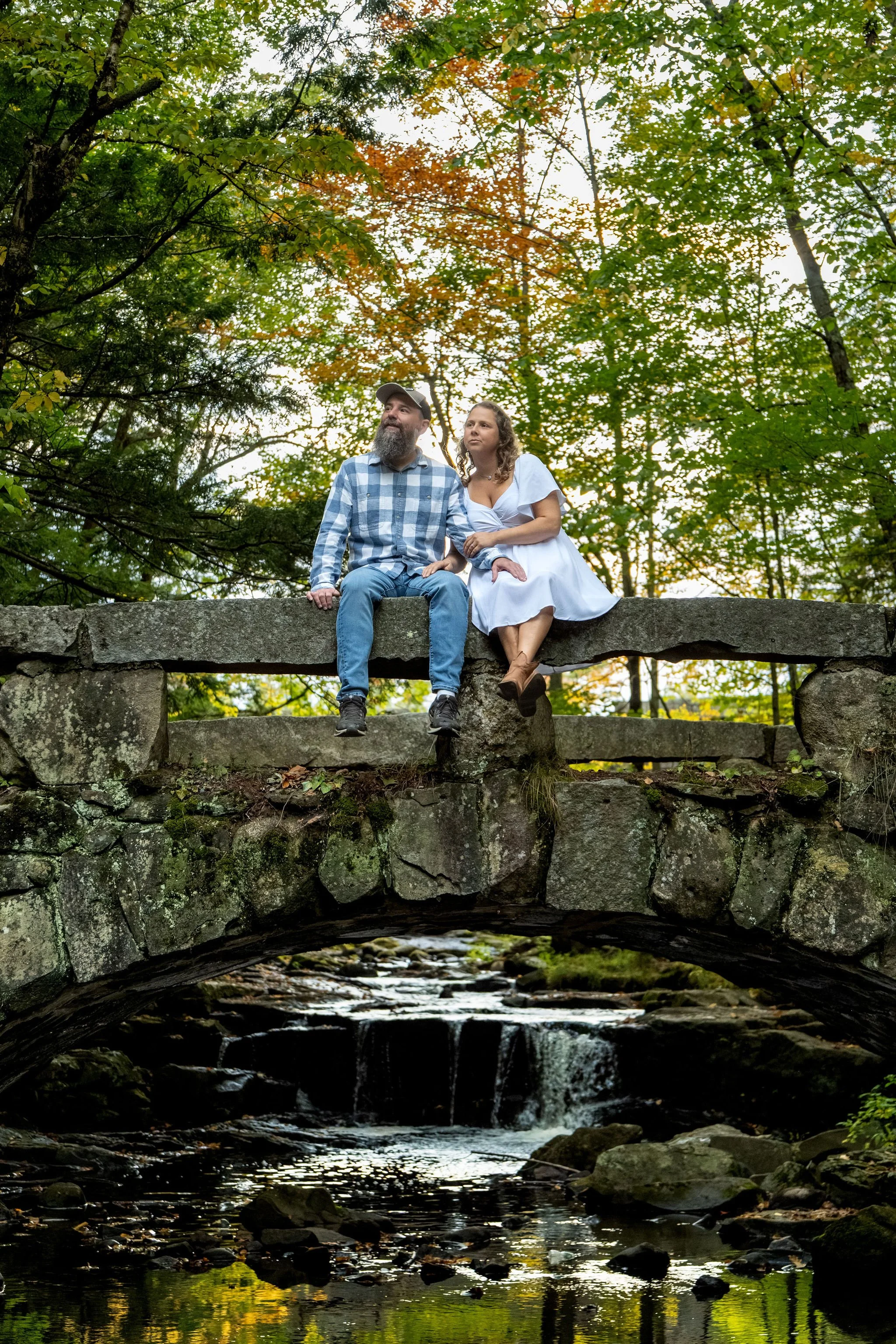 Alison and Dave looking at each other while sitting on the edge of the stone bridge at Vaughan Woods in Hallowell Maine fall engagement photography VMG Productions