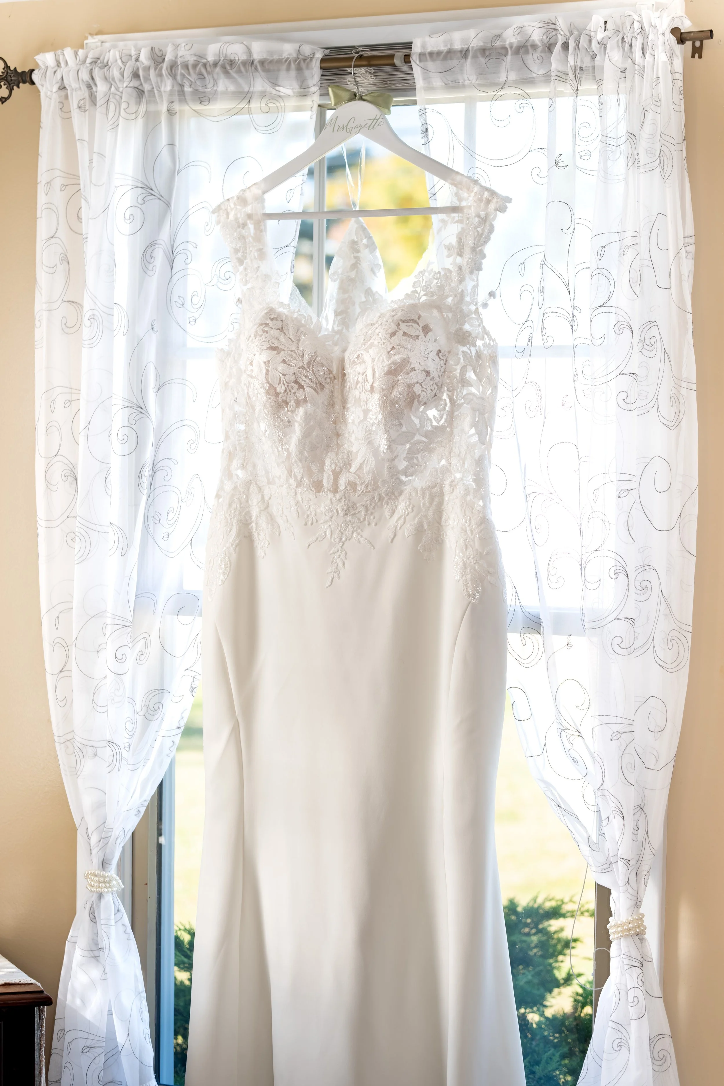 Erika's lace wedding dress hanging from a hanger in a sunny window with sheer curtains at the Maine Wedding Barn and Event Center in Minot Maine fall wedding photography VMG Productions