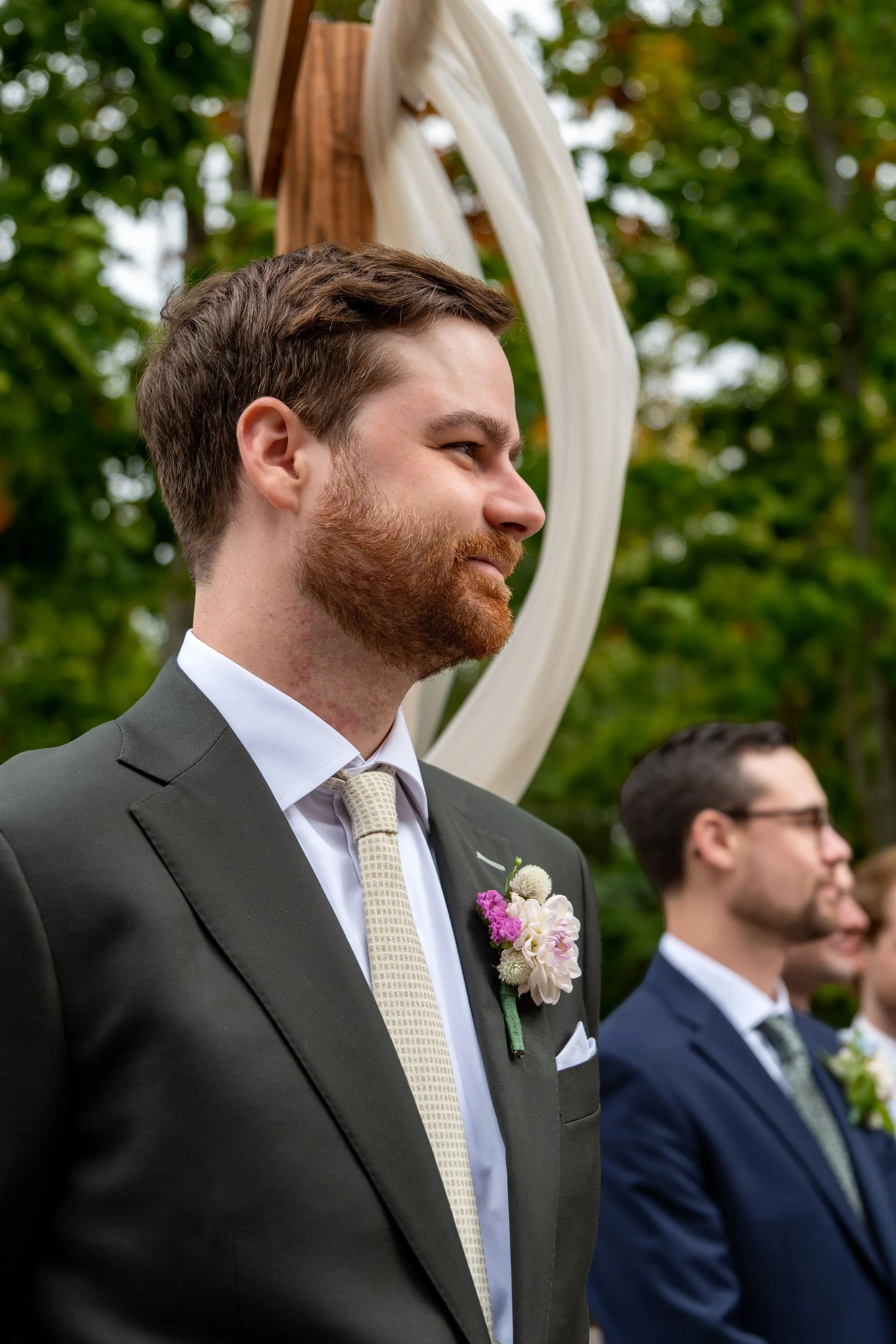 Jack watching Victoria come down the aisle with the white draped arbor behind him at The Cellars at Allagash Brewing Company in Portland Maine fall wedding photography VMG Productions