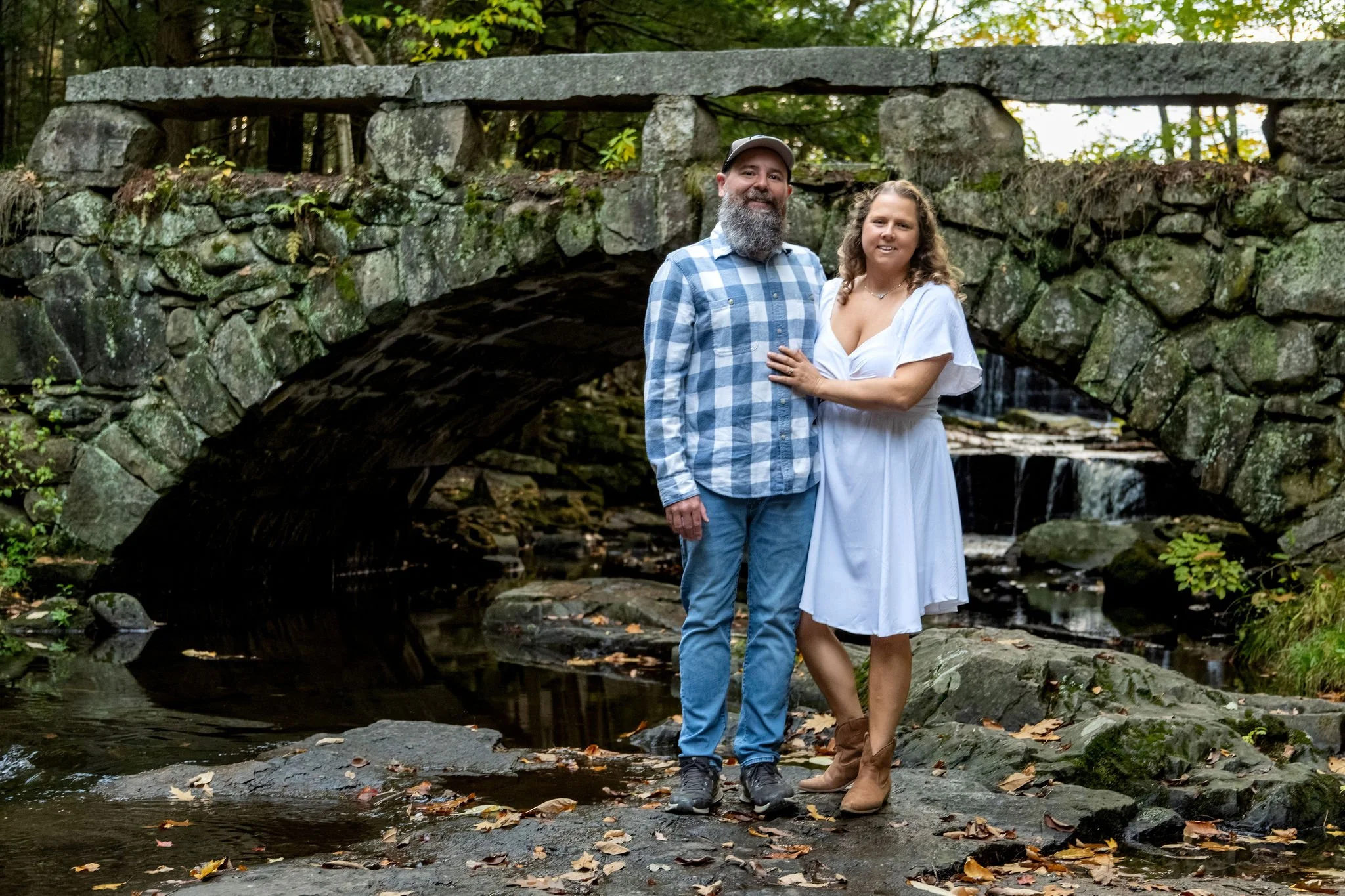 Alison and Dave standing arm in arm on the rocks in front of the granite arch bridge with the brook falling behind them at Vaughan Woods in Hallowell Maine fall engagement photography VMG Productions