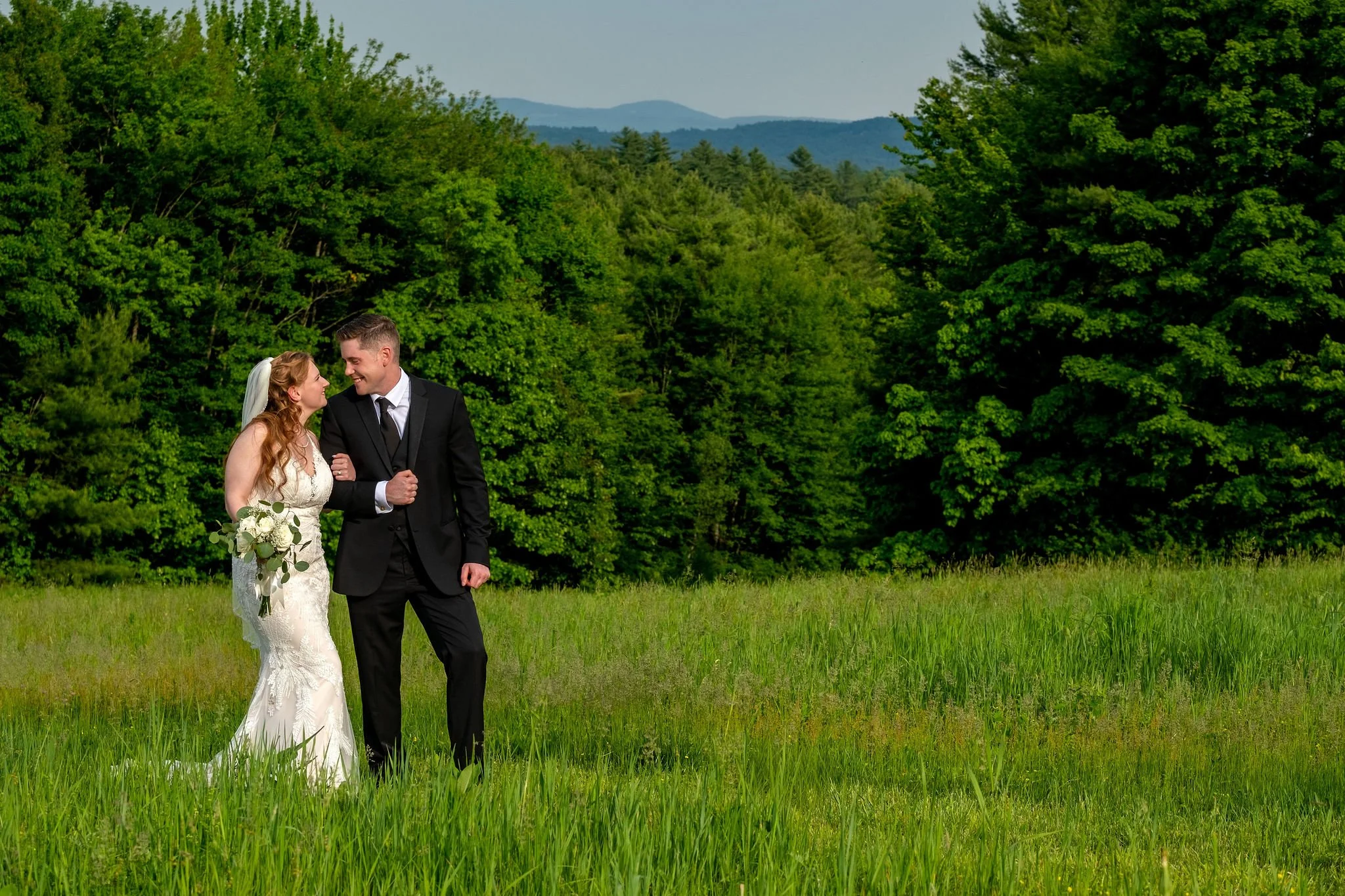 Kayla and Ethan DeAngelis walking together in the rolling field with the New Hampshire mountains behind them at their Sanbornton wedding photography VMG Productions