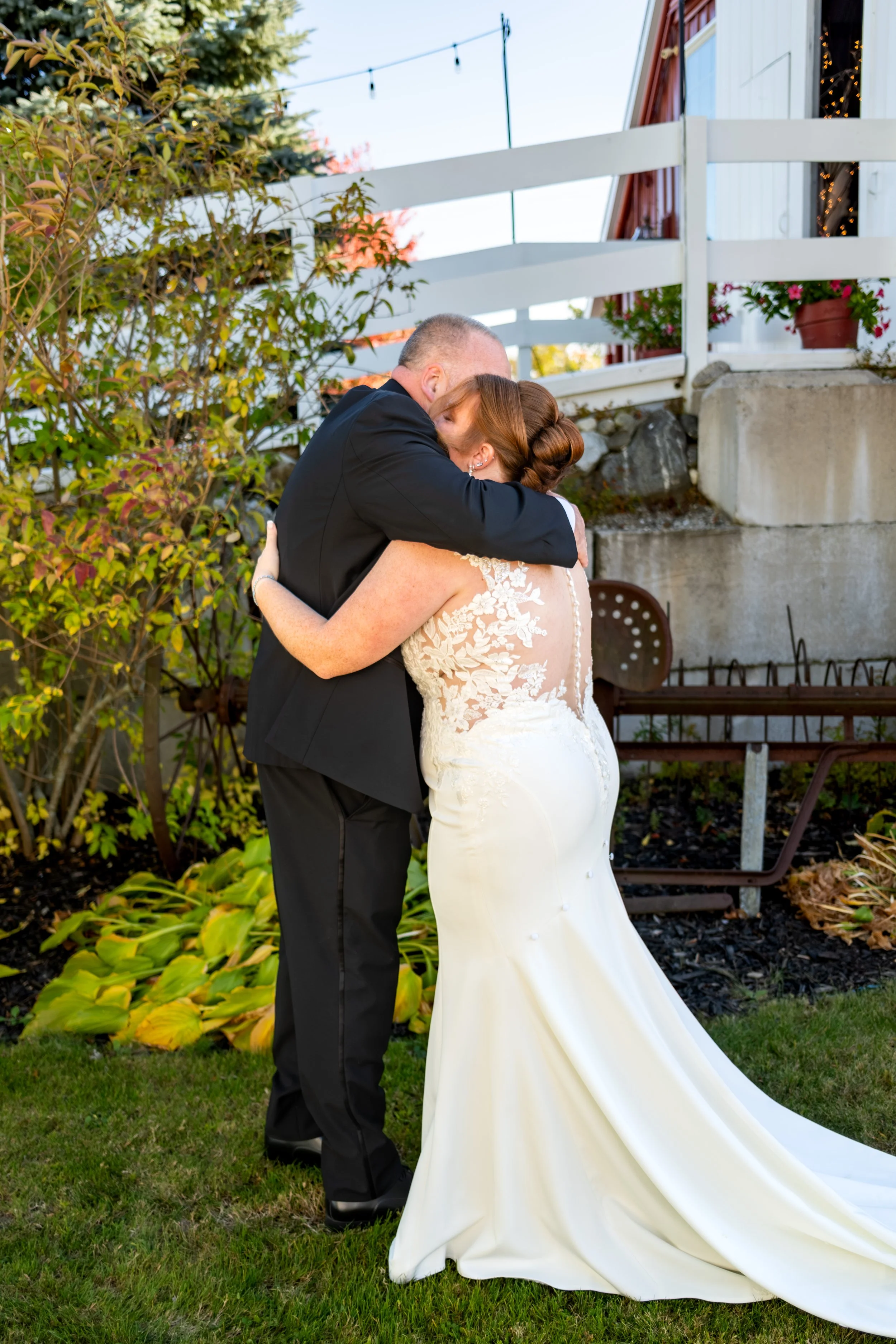 Erika hugging her dad in front of the white fence and red barn at the Maine Wedding Barn and Event Center in Minot Maine fall wedding photography VMG Productions