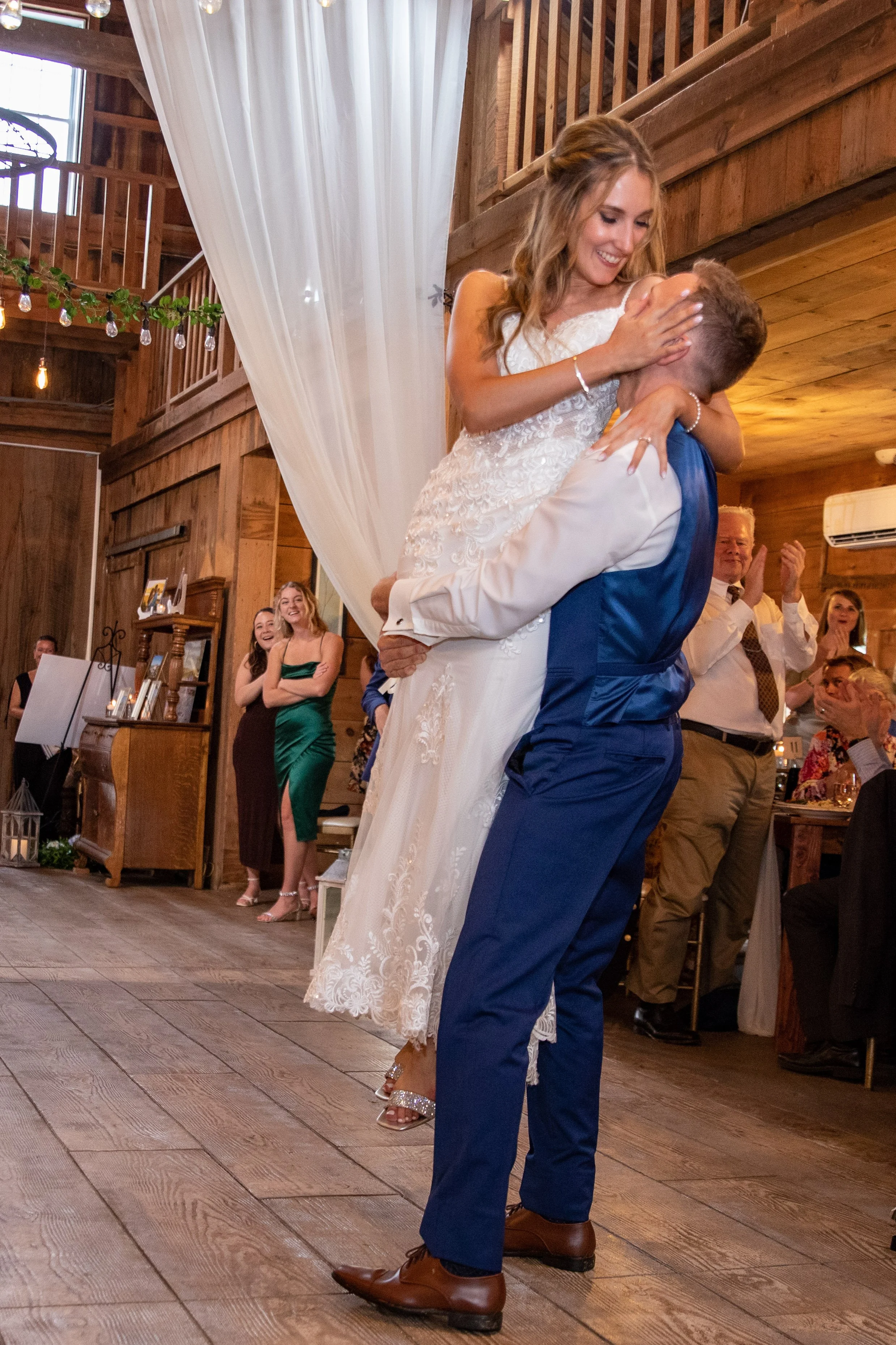 Groom in navy blue suit lifting smiling bride in white lace wedding gown during first dance at River Winds Farm and Estate Saco Maine 2023 wedding with guests applauding and laughing in background inside barn venue with string lights and white fabric drape