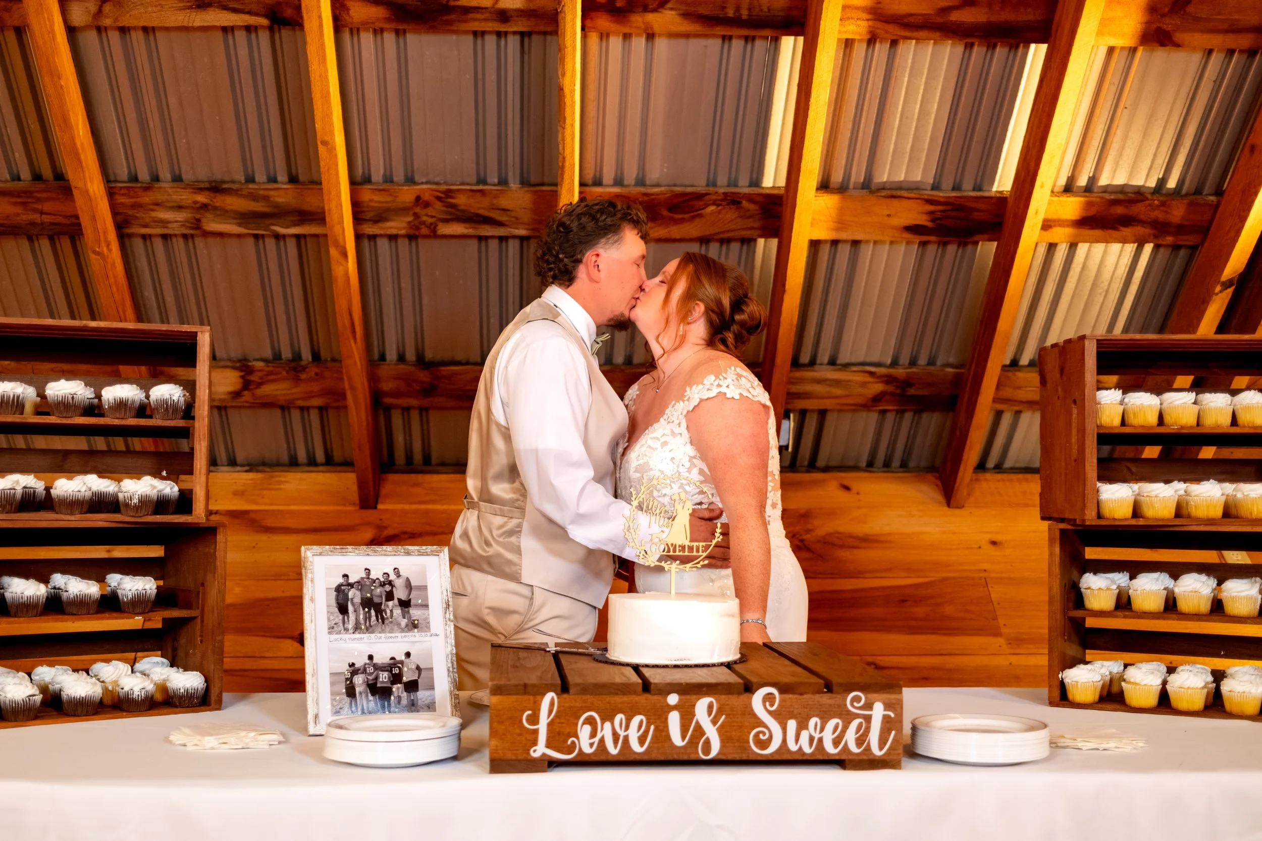 Andrew and Erika kissing over their cake with a cupcake display behind them at the Maine Wedding Barn and Event Center in Minot Maine fall wedding photography VMG Productions