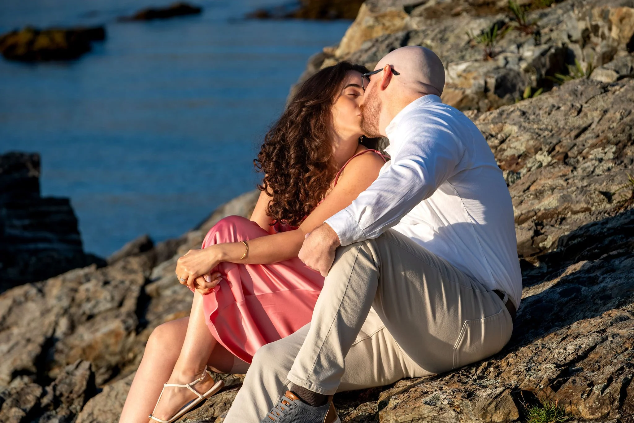 Zack and Helena kissing as they sit together on the rocks above the water on Marginal Way in Ogunquit Maine sunrise proposal and engagement photography VMG Productions