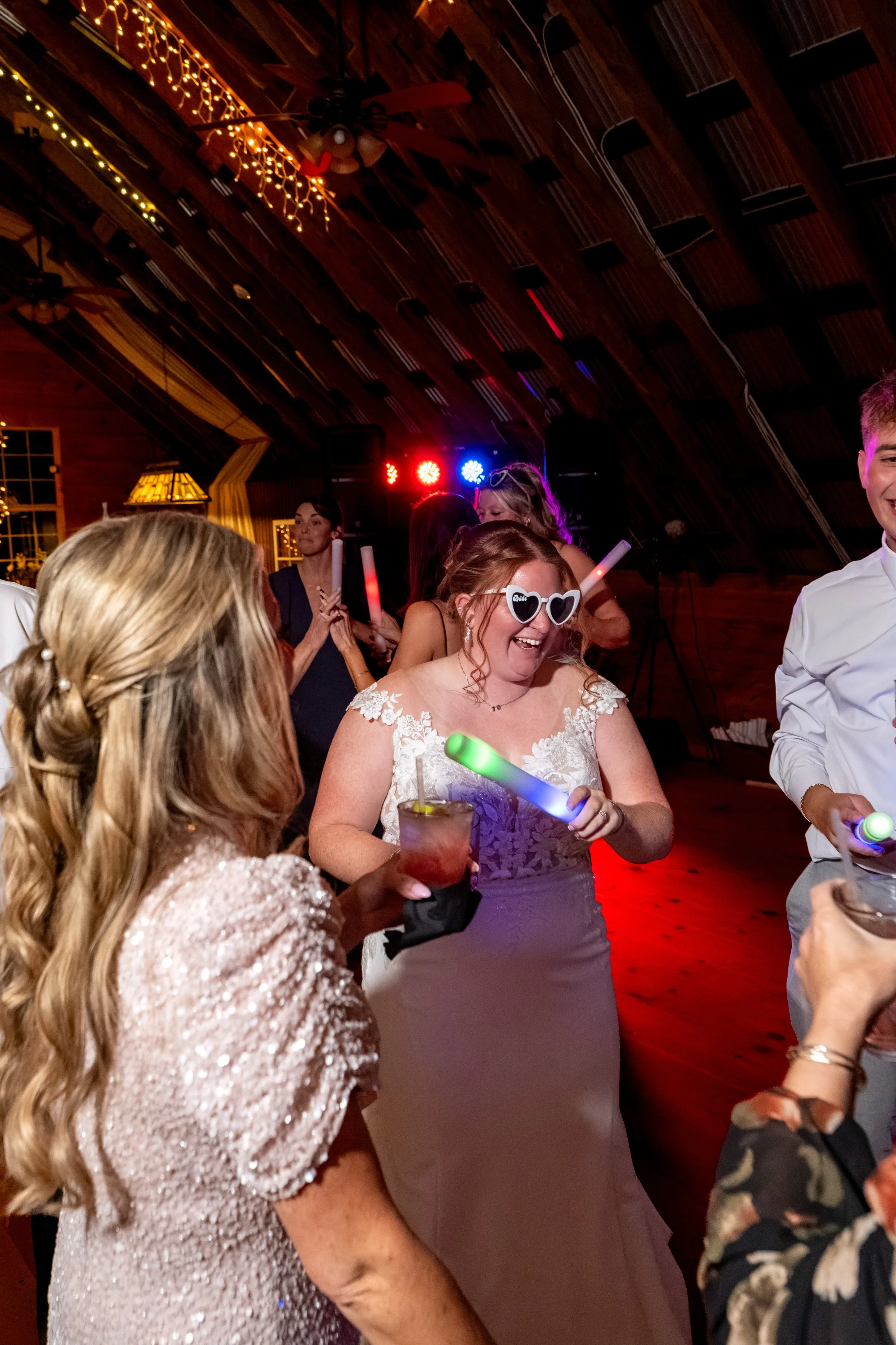 Erika in white heart-shaped sunglasses dancing with a wand in her hand as friends dance around her at the Maine Wedding Barn and Event Center in Minot Maine fall wedding photography VMG Productions