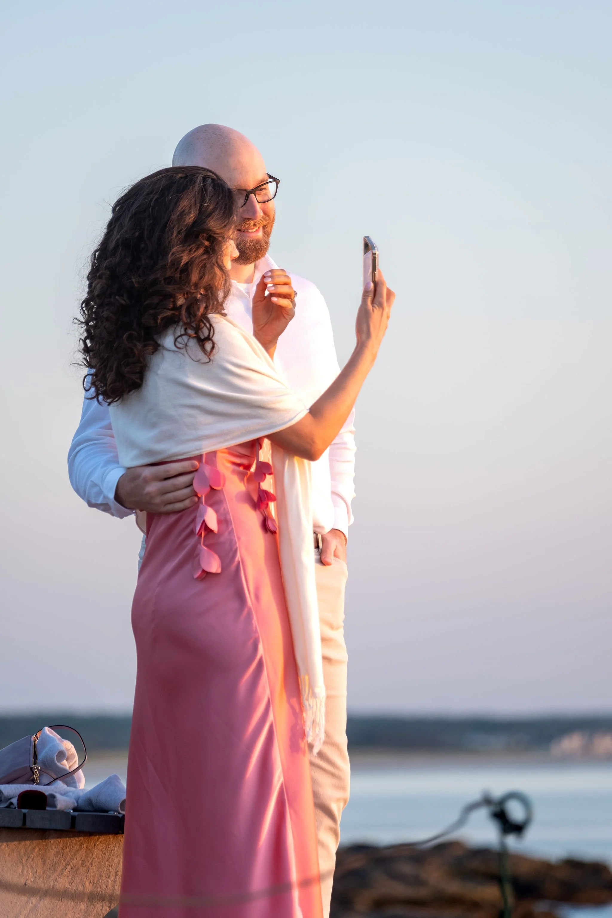 Helena holding up her phone to take a selfie with Zack, wrapped in a cream shawl over her pink dress on Marginal Way in Ogunquit Maine sunrise proposal and engagement photography VMG Productions