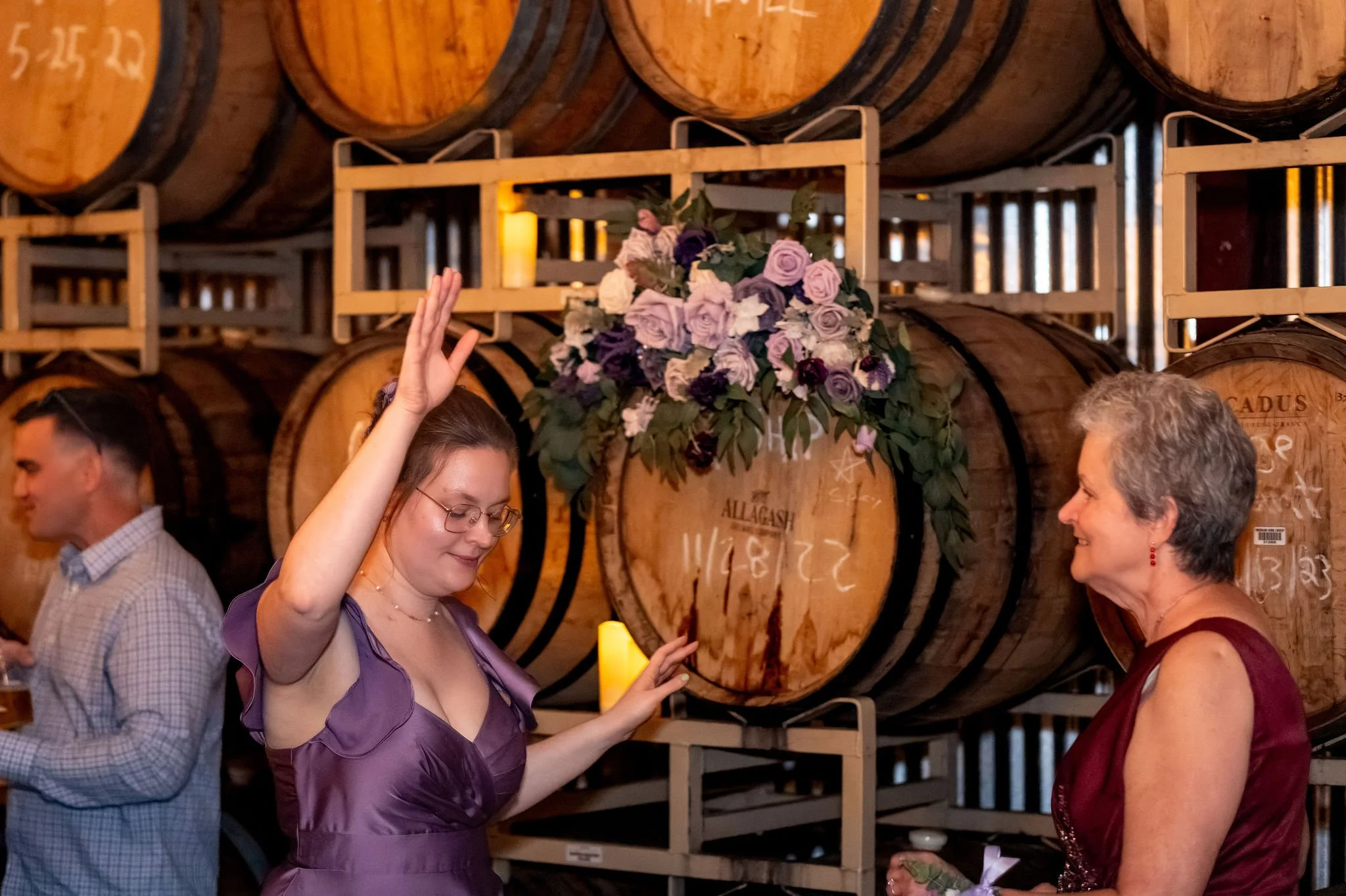 A bridesmaid in lavender dancing with her arm in the air beside a wreath of purple roses on a barrel at The Cellars at Allagash Brewing Company in Portland Maine fall wedding photography VMG Productions
