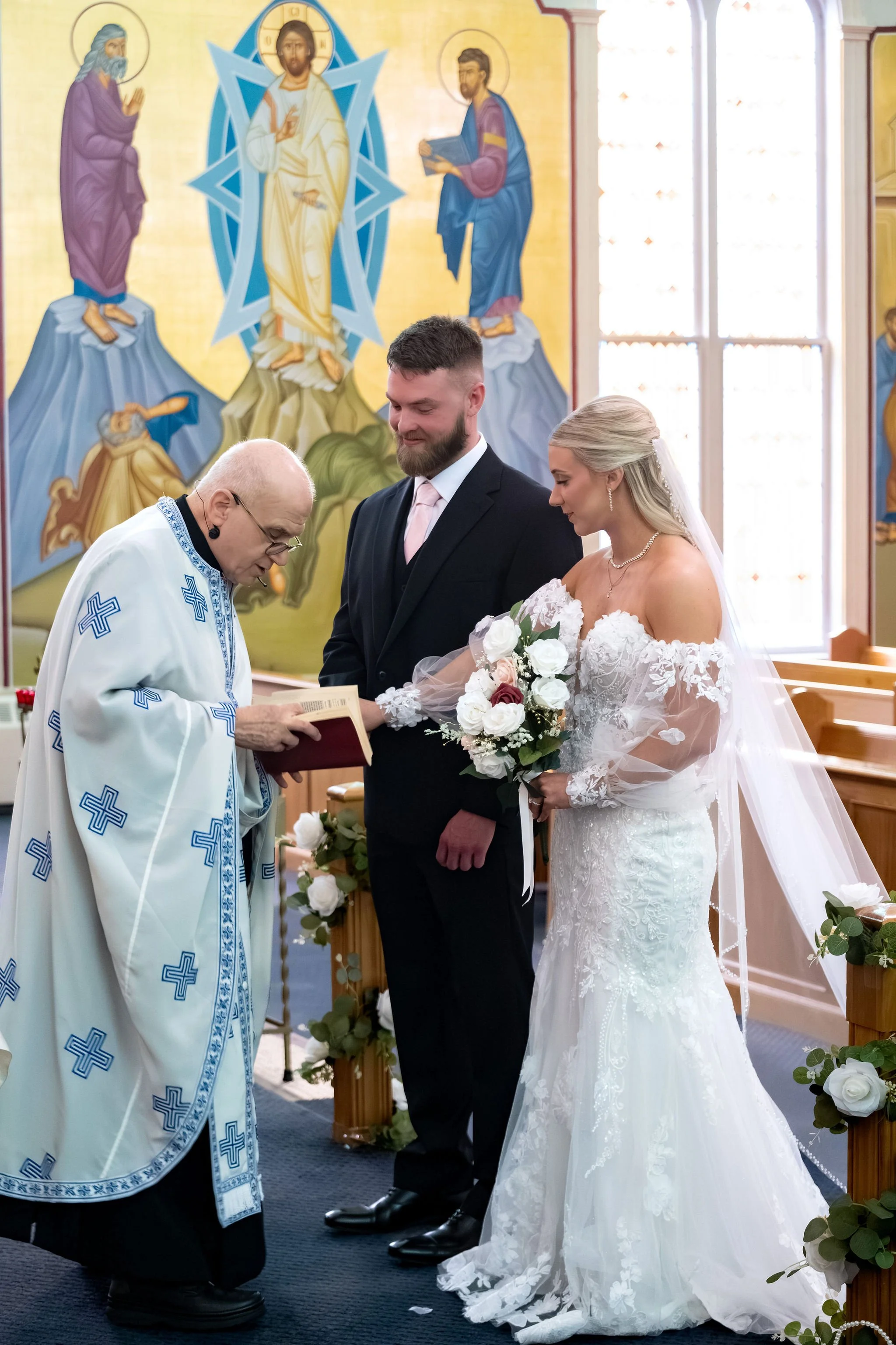 Jeremiah and Vienna stand together while the priest performs part of the ceremony at Holy Trinity Greek Orthodox Church Portland Maine spring wedding photography VMG Productions