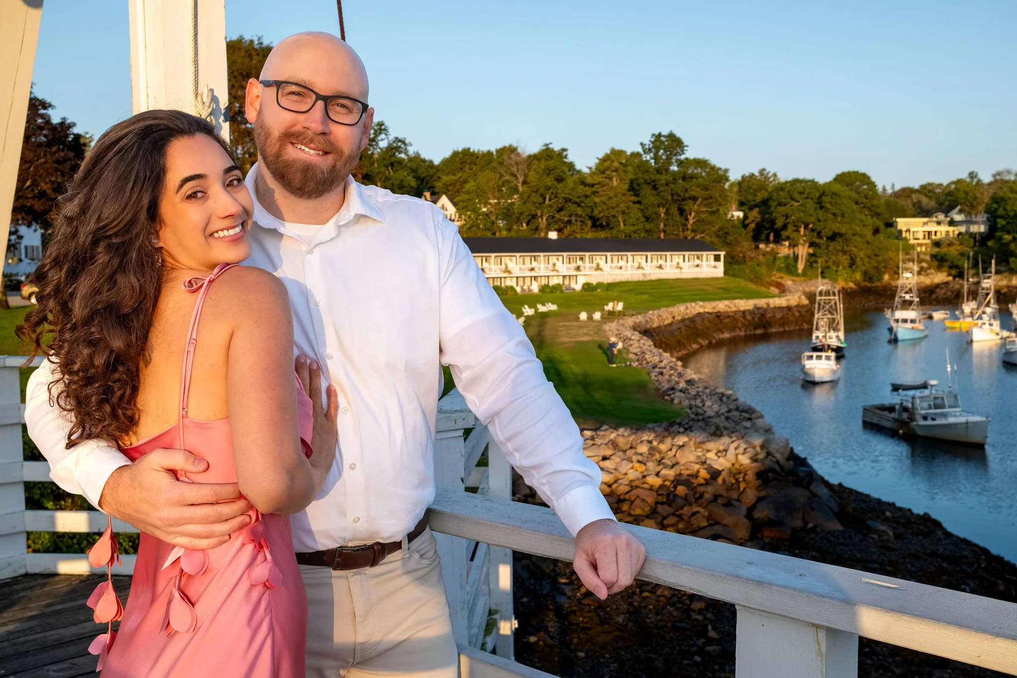 Helena smiling over her shoulder as Zack holds her from behind at the white railing of the footbridge with the harbor below on Marginal Way in Ogunquit Maine sunrise proposal and engagement photography VMG Productions