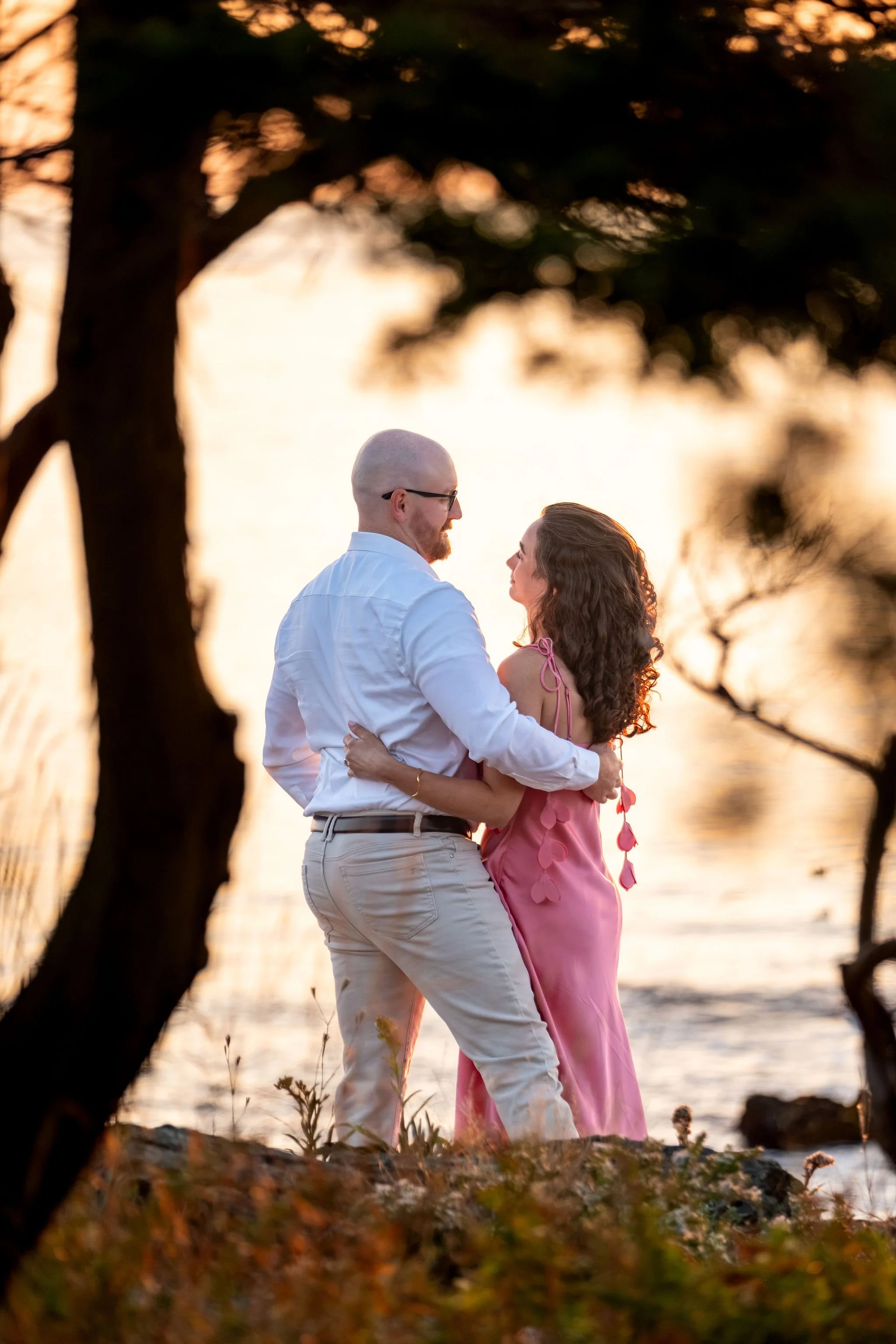 Zack and Helena standing arm in arm looking out at the ocean through the branches of a tree on Marginal Way in Ogunquit Maine sunrise proposal and engagement photography VMG Productions