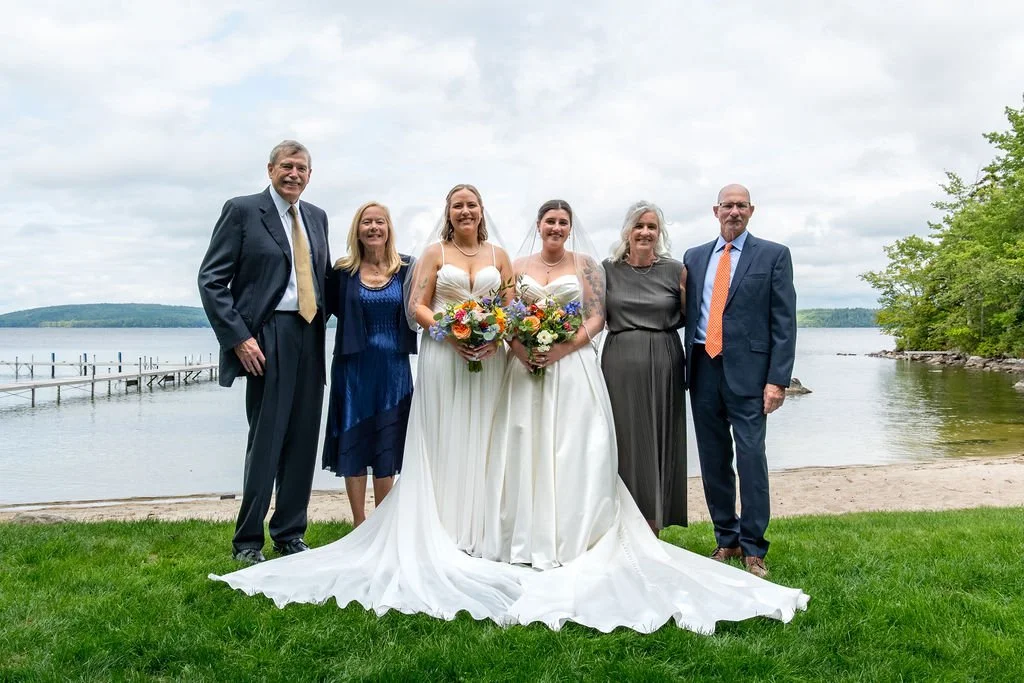 Two brides Emma and Hannah in white wedding gowns with both sets of parents formal family portrait on grassy waterfront lawn with dock and lake in background at Camp Runoia Belgrade Maine summer 2025 wedding