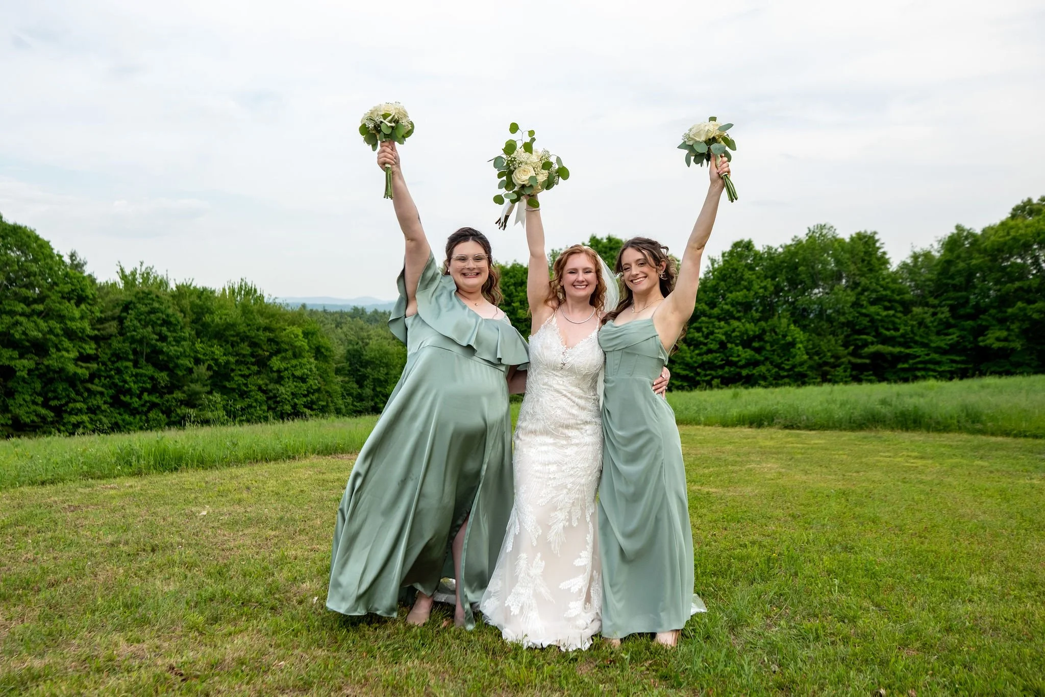 Kayla DeAngelis and her bridesmaids raising their bouquets in the summer field at their Sanbornton New Hampshire wedding photography VMG Productions