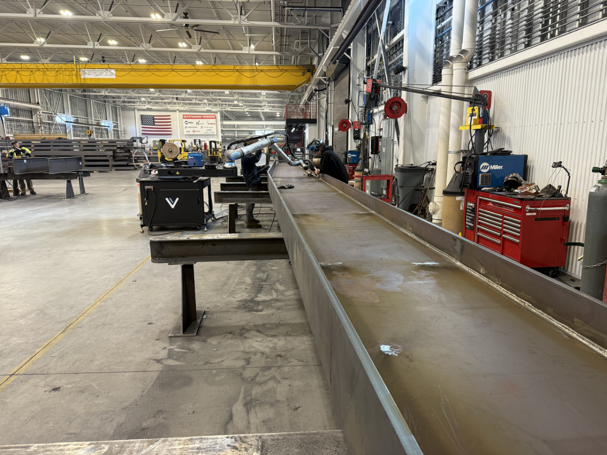 Structural steel facility with a robotic welding arm working on a large steel beam, demonstrating automated structural steel manufacturing and precision fabrication processes.