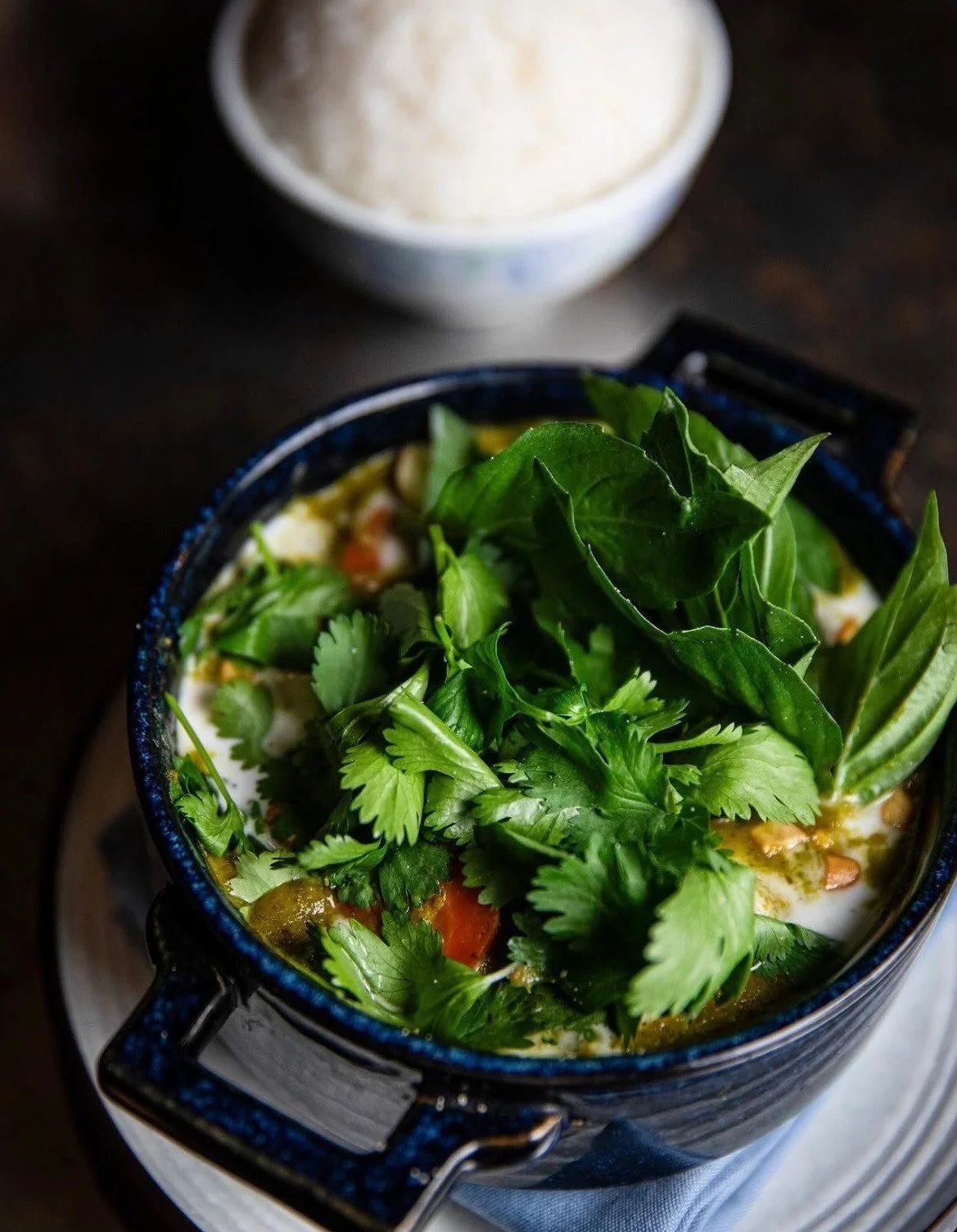A blue handled bowl of vegetables and sauce topped with fresh herbs, with rice behind it.