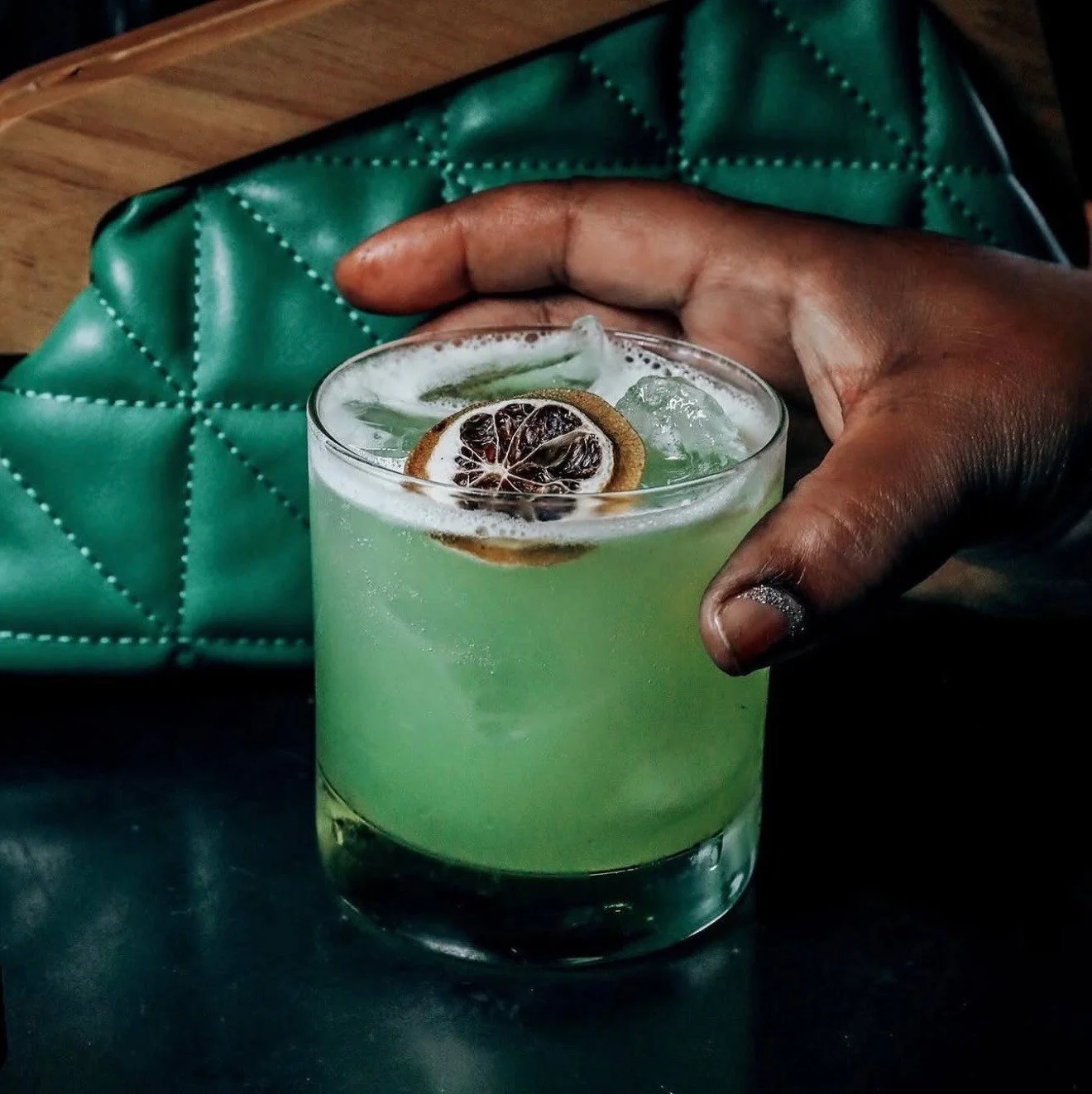 A hand holding a green iced drink garnished with a dried citrus slice.