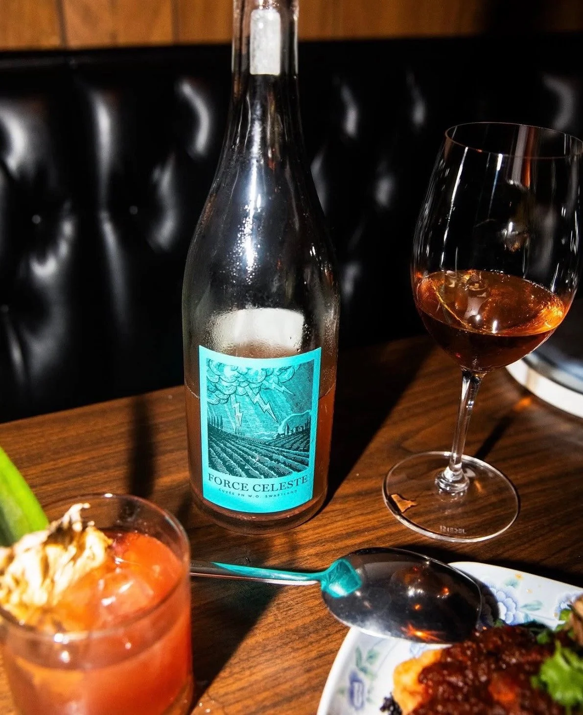 A wine bottle and glass beside an orange drink and plated food on a wooden table.
