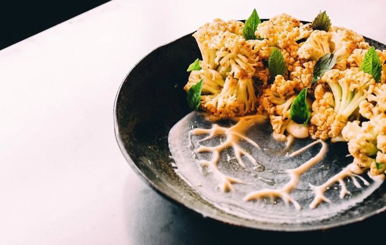 Cauliflower florets with fresh herbs and a branching sauce pattern on a dark plate.