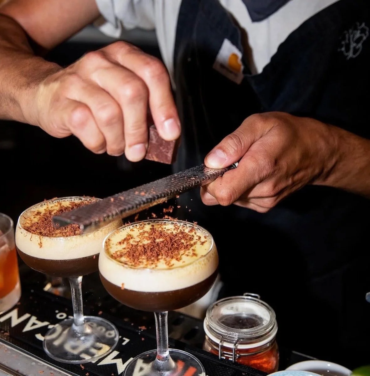 Hands grating chocolate over two dark drinks with creamy foam in coupe glasses.