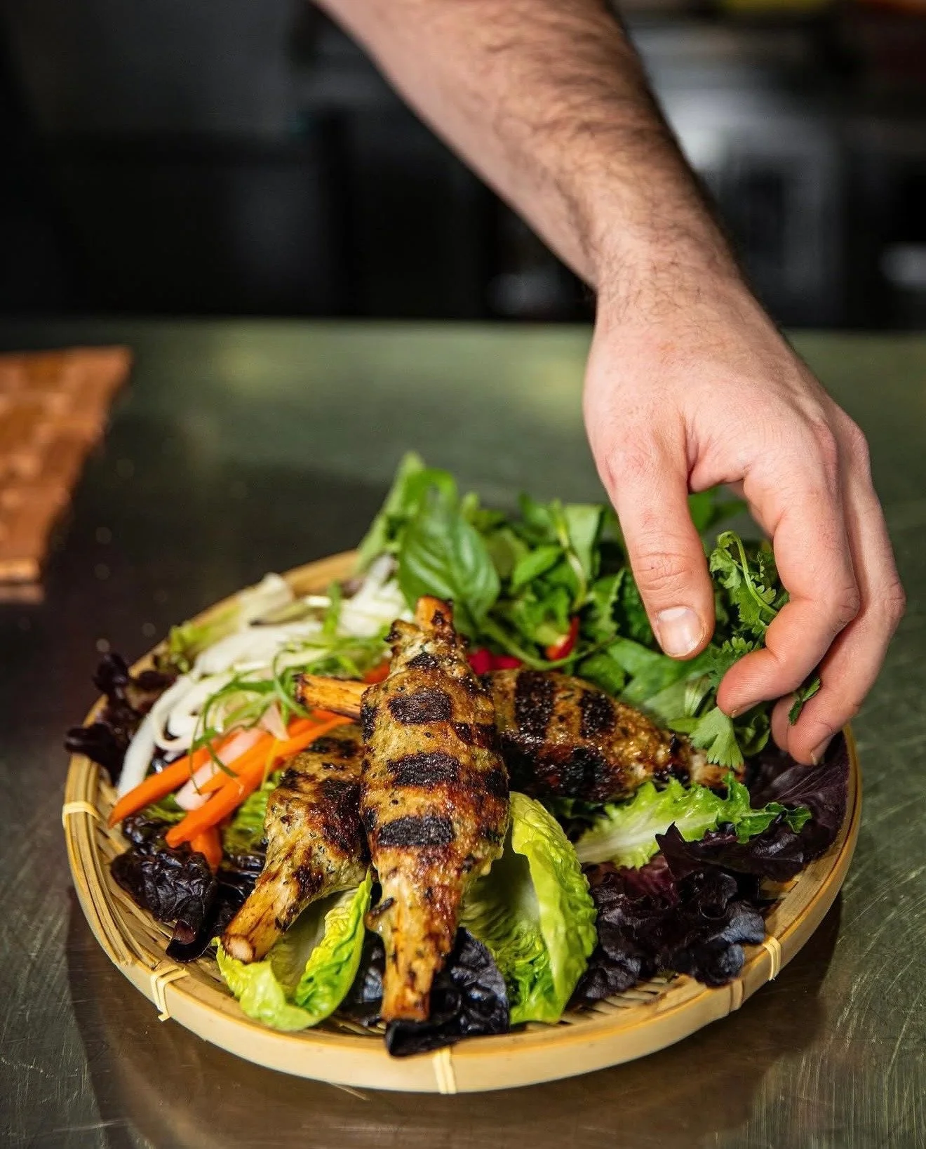 A hand arranging herbs beside grilled skewers on a woven serving tray.