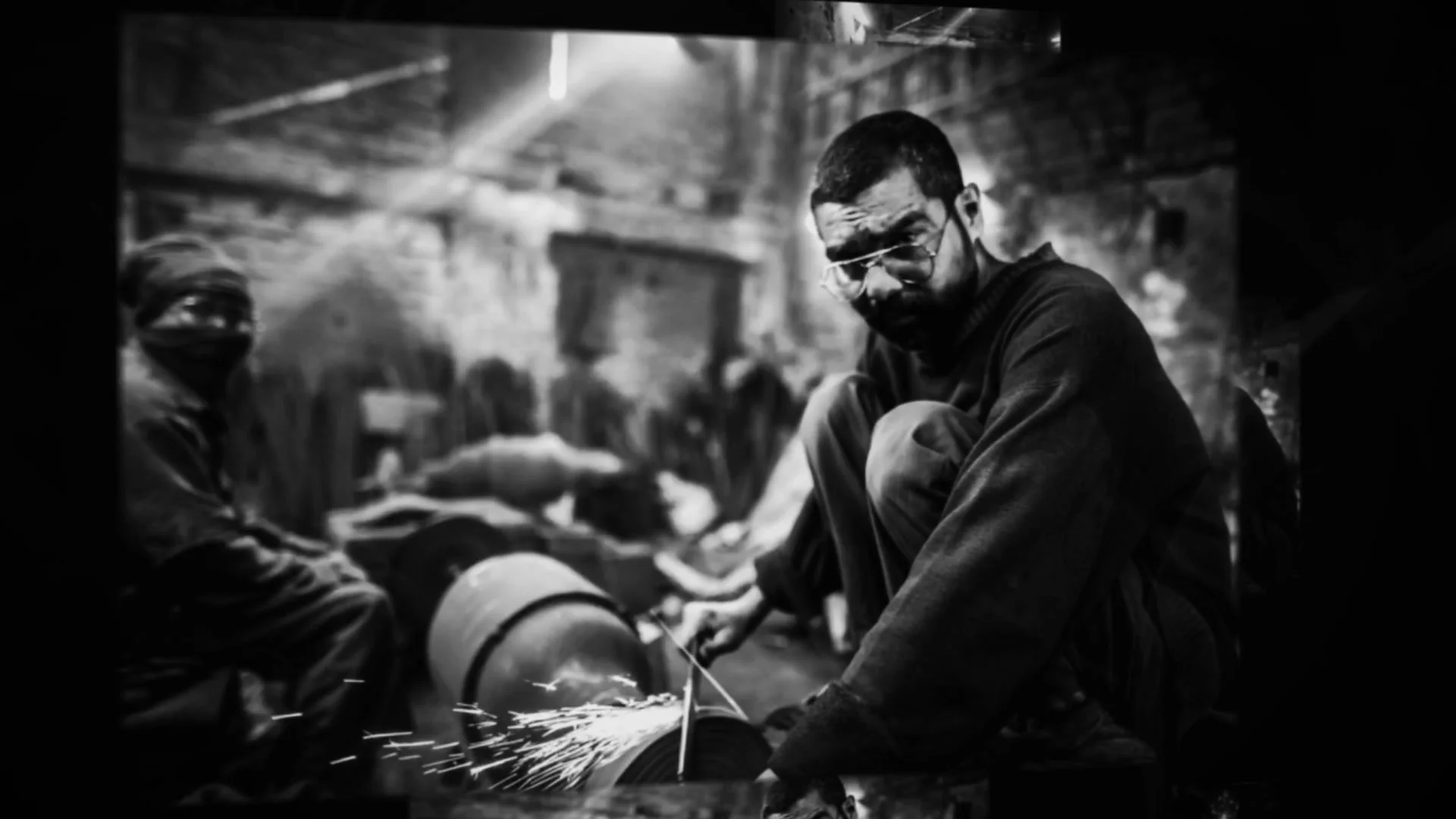 A karigar grinds a blade at the wheel, sparks flying, in black and white