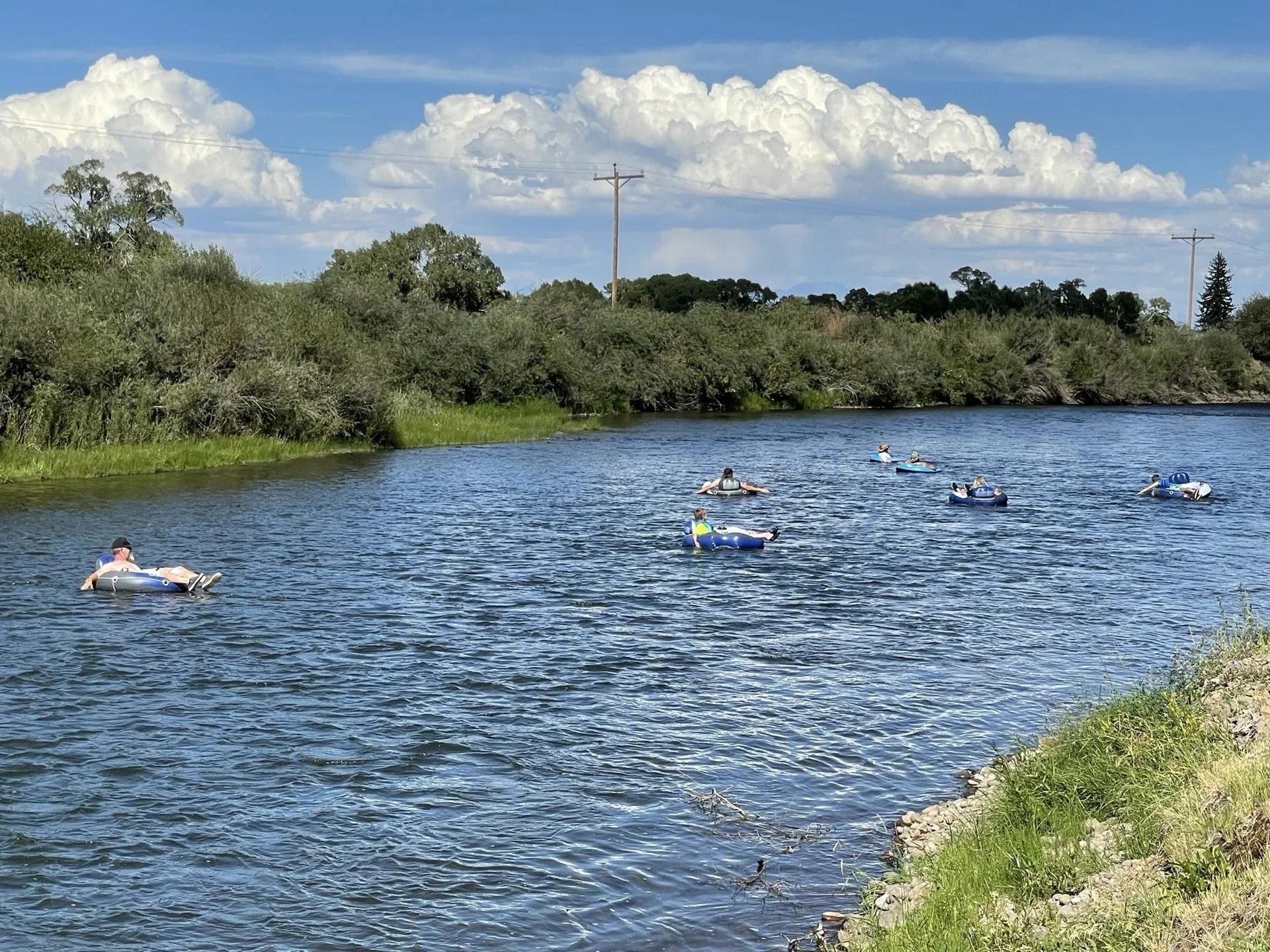 Guests tubing on the Rio Grande beneath a sunny blue sky, beside green riverbanks.