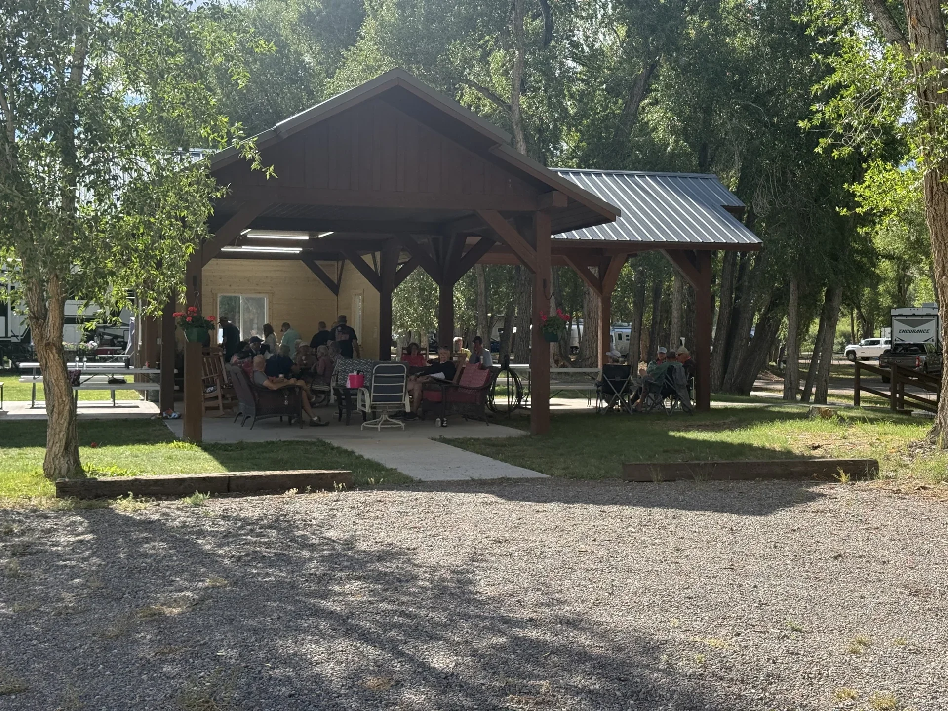Guests gathered beneath the wooden pavilion at Woods and River RV Park, surrounded by shade trees.