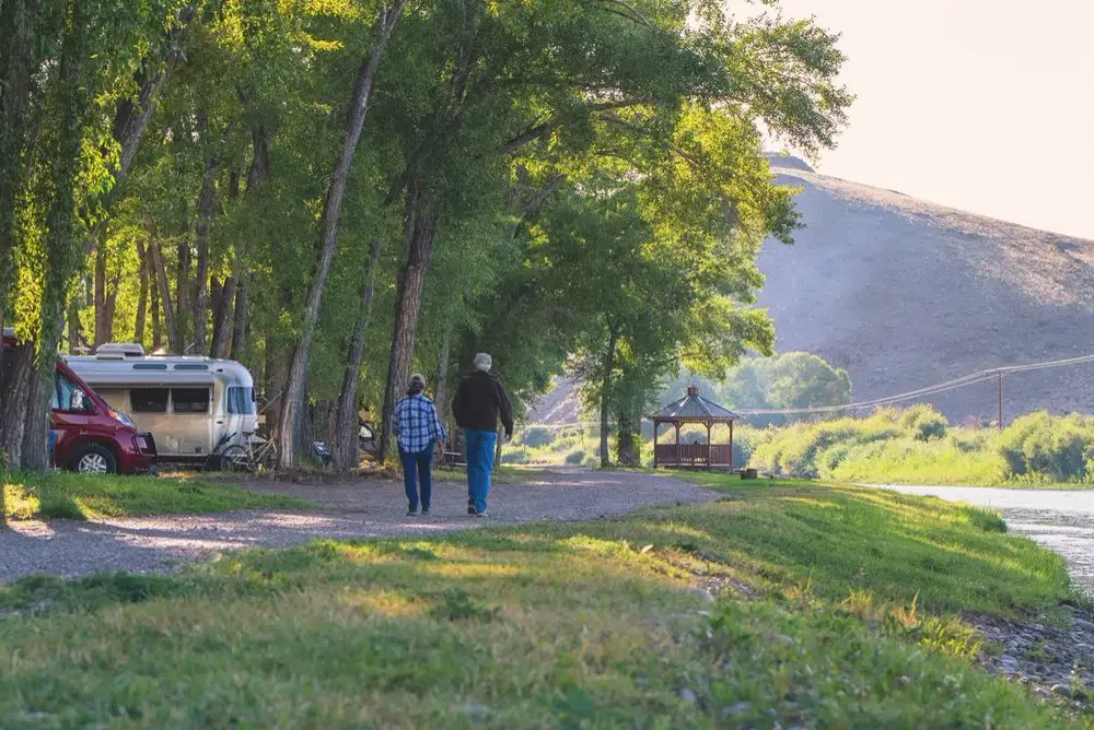 Two people walking beneath shade trees beside RV campsites and the river.